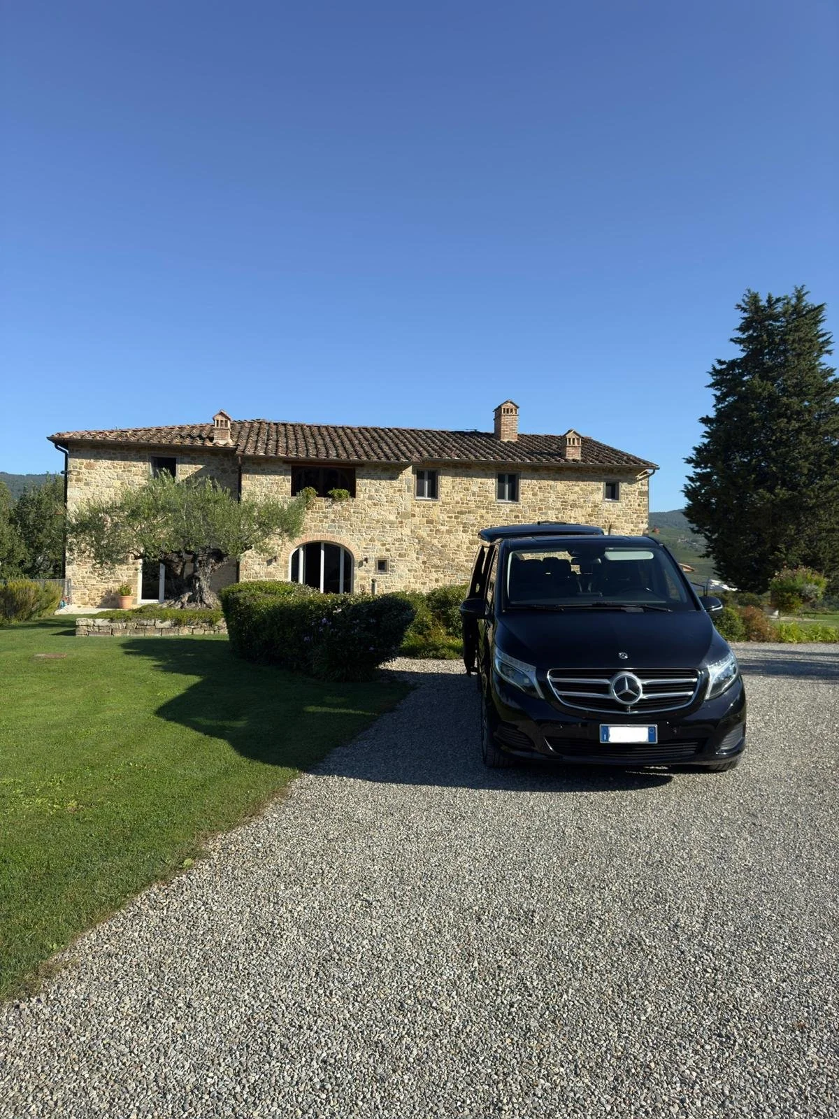 Una casa in pietra con un tetto di tegole, un albero di ulivo e un'auto Mercedes nera parcheggiata nel cortile.