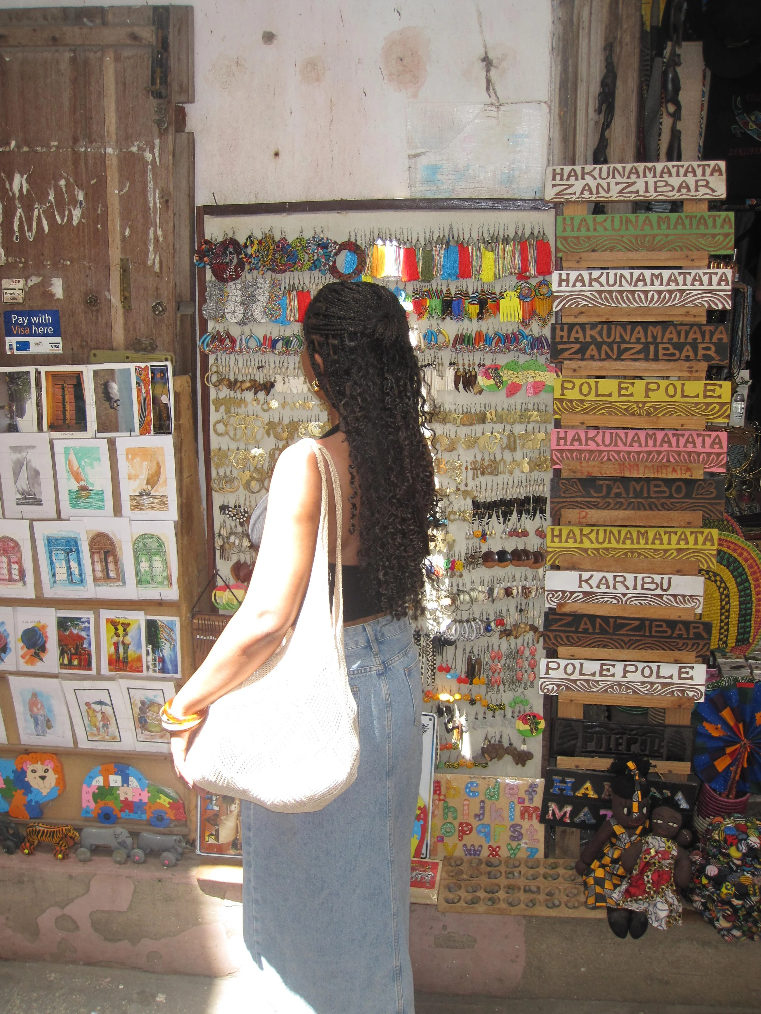 Woman with long curly hair shopping at a colorful souvenir stand displaying jewelry, wooden signs, and handmade crafts in a market.