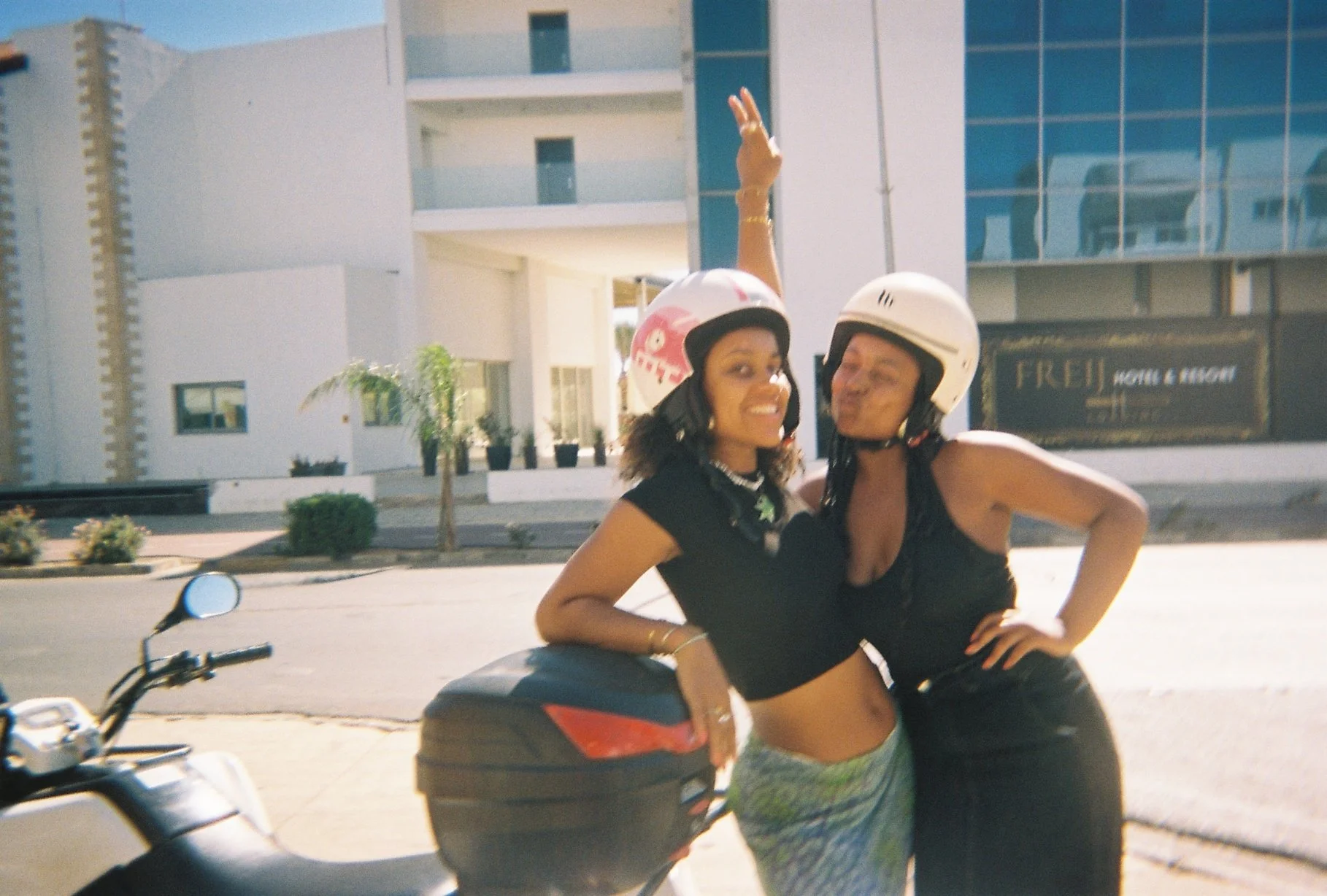 Two women wearing helmets are standing next to a scooter on a city street, smiling and posing for the camera.