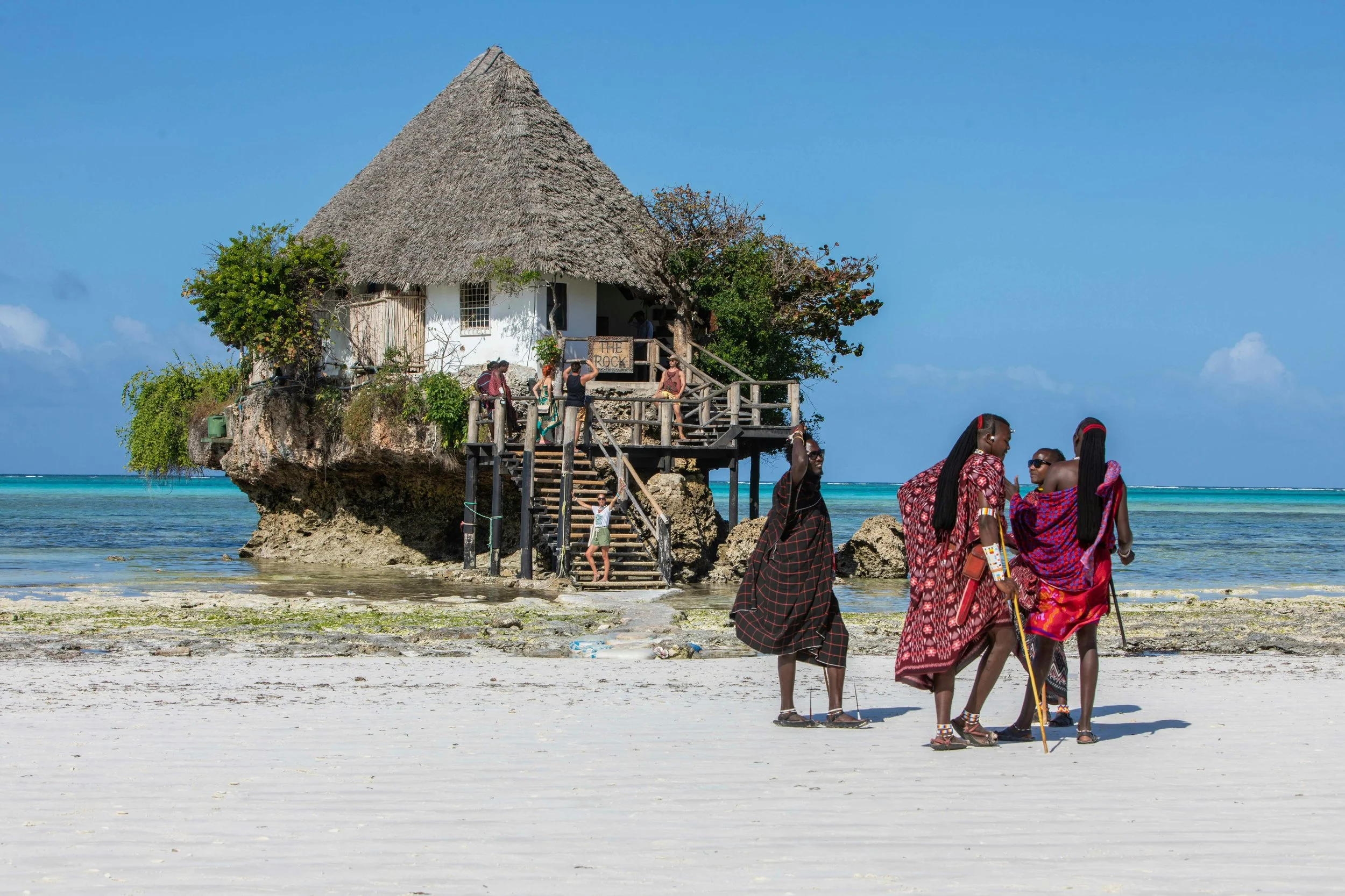 A thatched-roof house on stilts over water with people in traditional Maasai attire walking on a sandy beach in front.