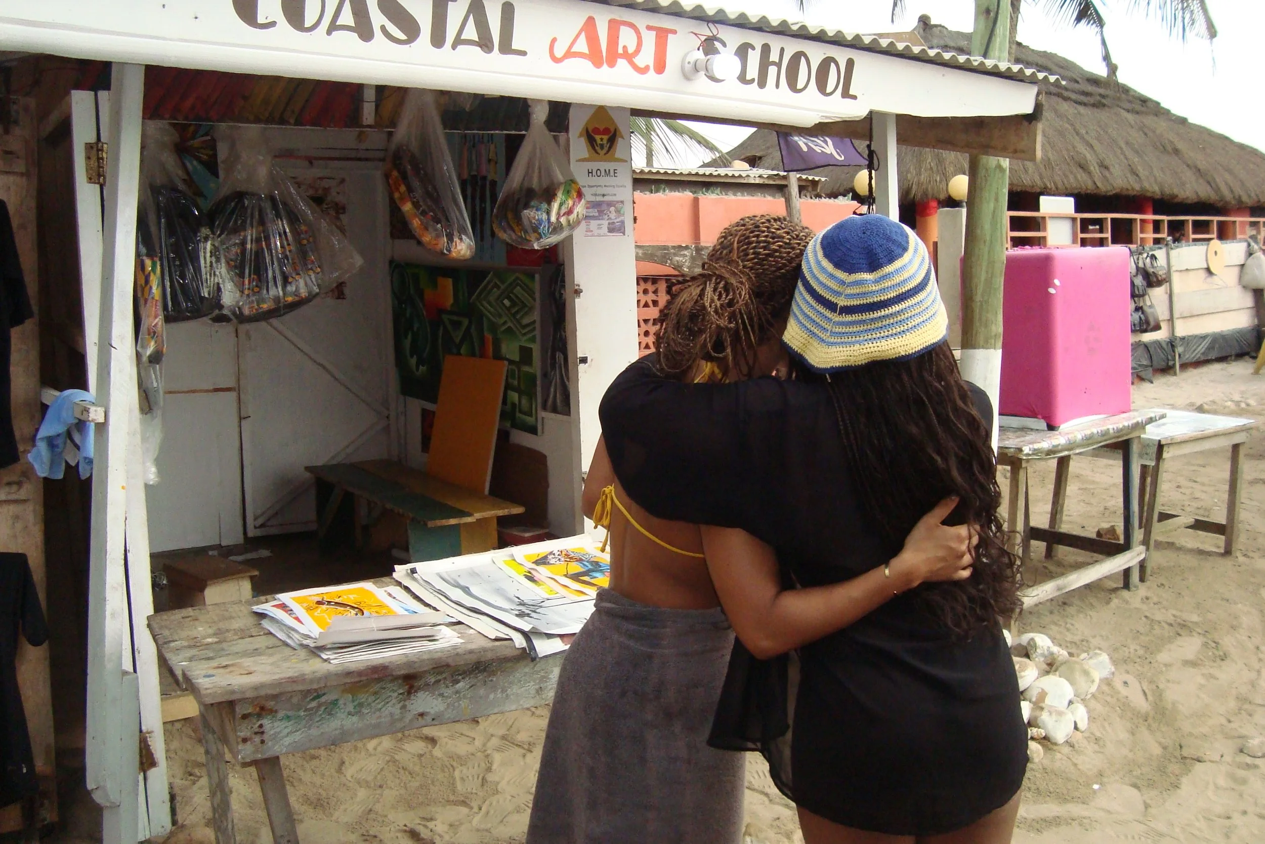 Two women hugging at a beachside art shop with paintings and bags displayed behind them.