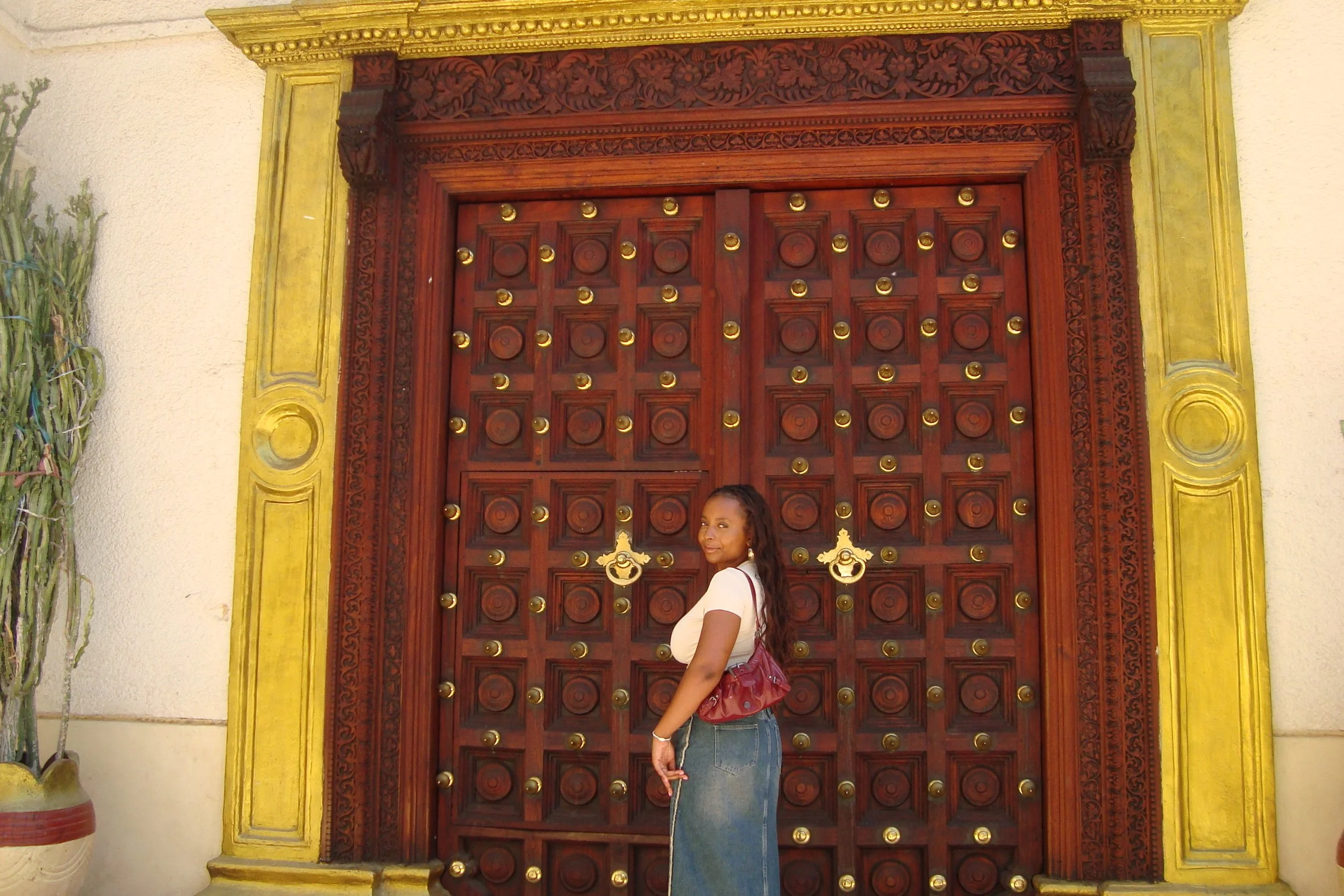 A woman with long, curly hair wearing a white top, blue jeans, and a red handbag standing in front of a large, ornate wooden door with gold accents and intricate carvings.