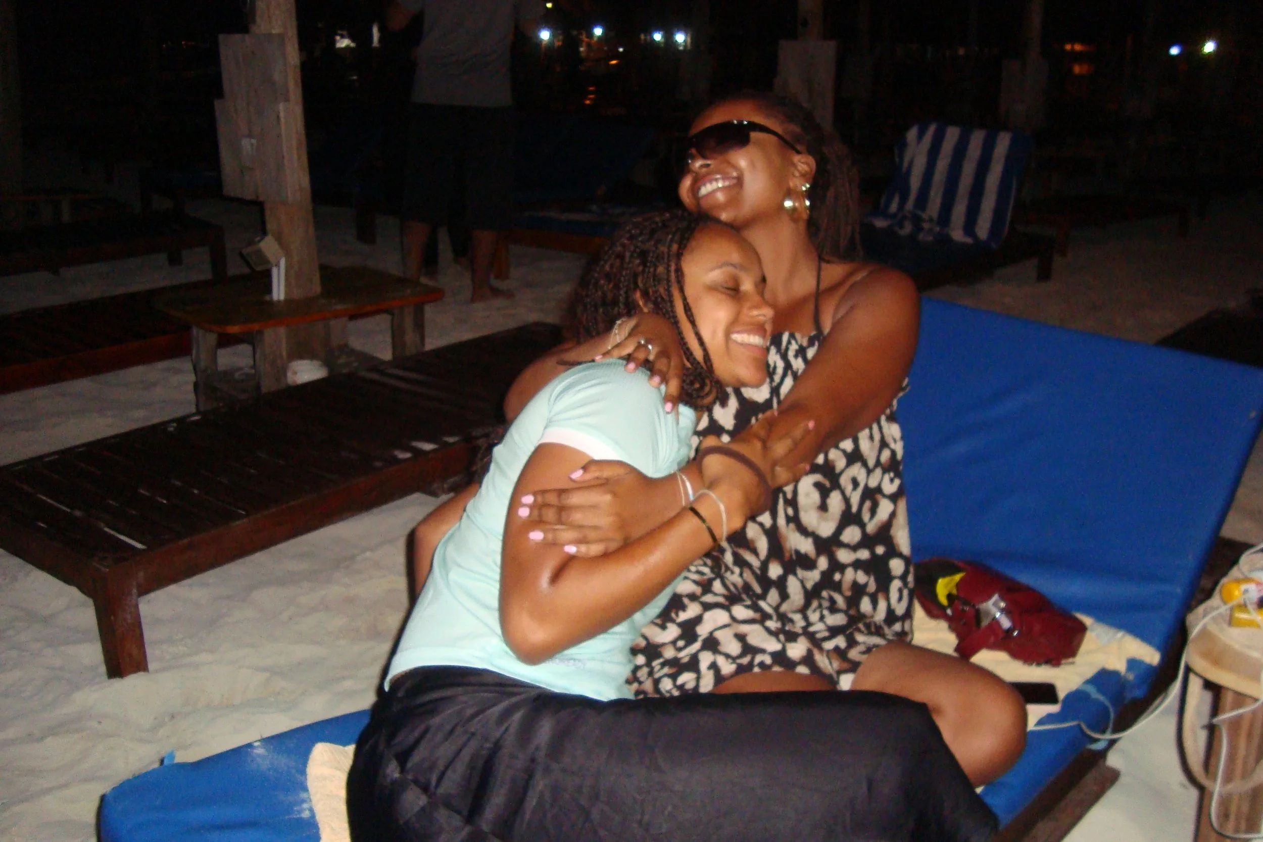 Two women are hugging and smiling at a beach lounge chair during nighttime, embracing each other happily.