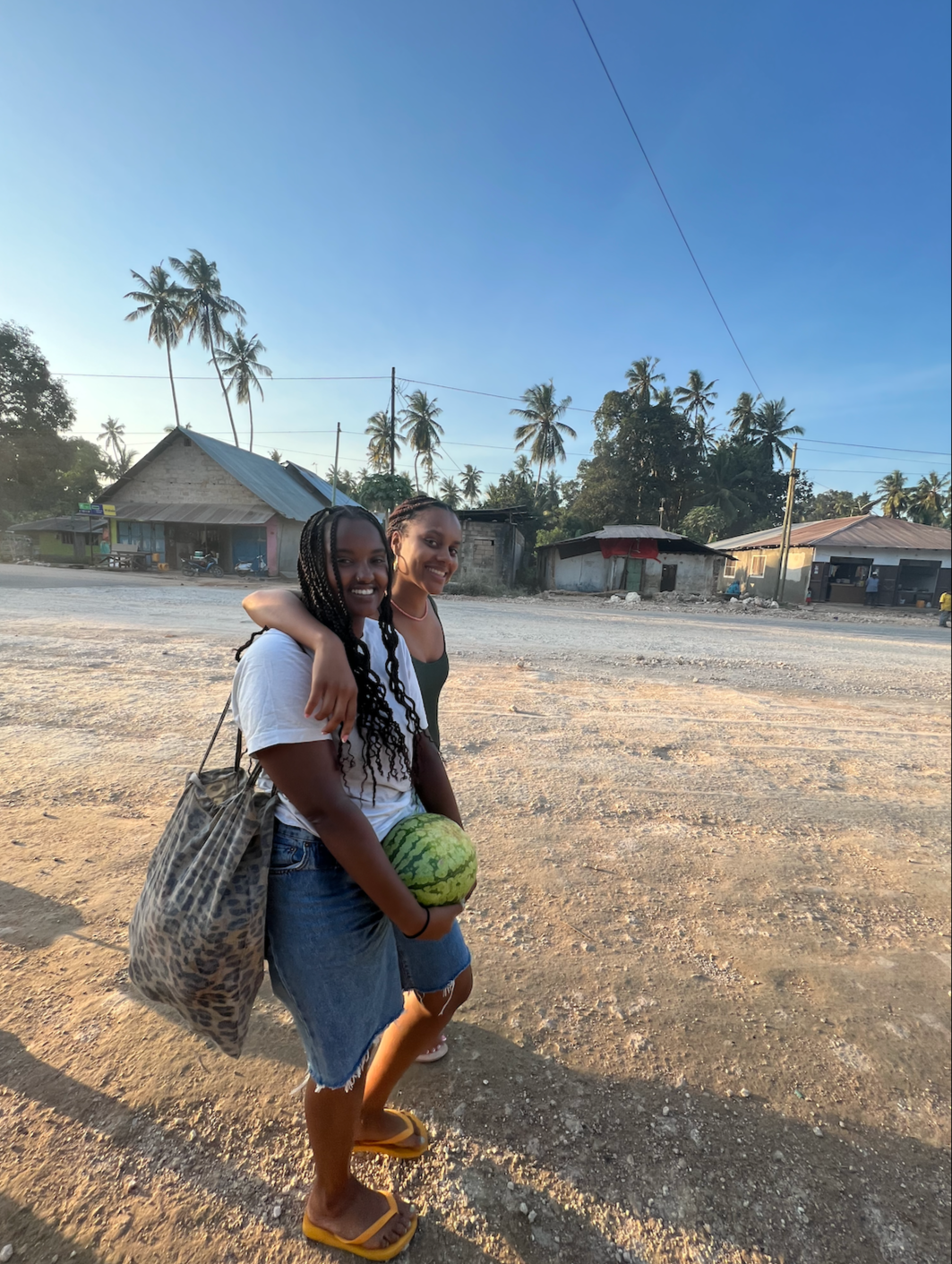 Two women smiling and hugging each other while holding a watermelon on a dirt road, with palm trees and simple houses in the background during sunset.