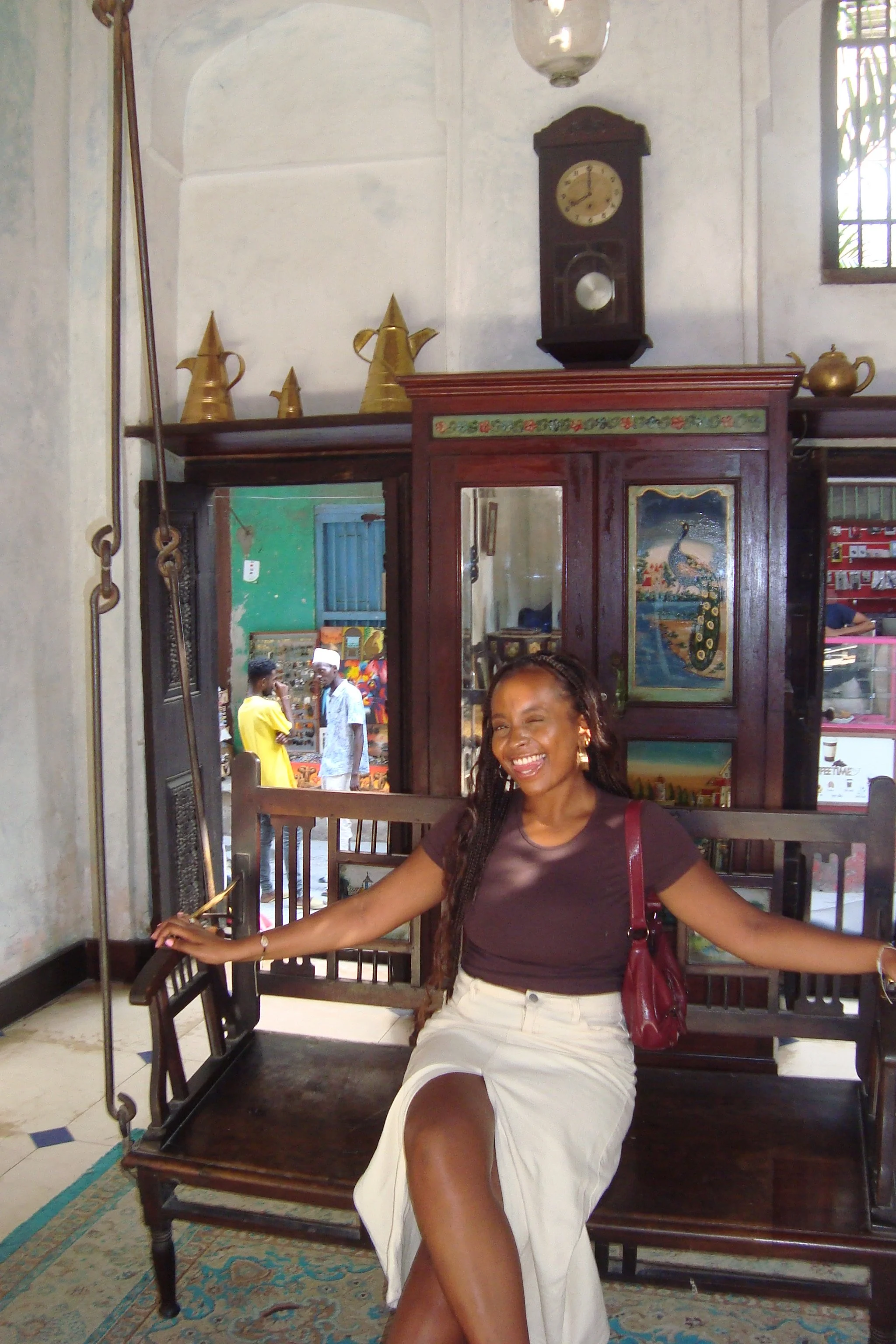 A woman with long braids, wearing a dark brown t-shirt and cream skirt, sitting on a wooden bench inside a room with vintage decor. She is smiling and extending her arms, with a handbag on her shoulder. Behind her is a mirror, a clock, and decorative items on a wooden shelf.