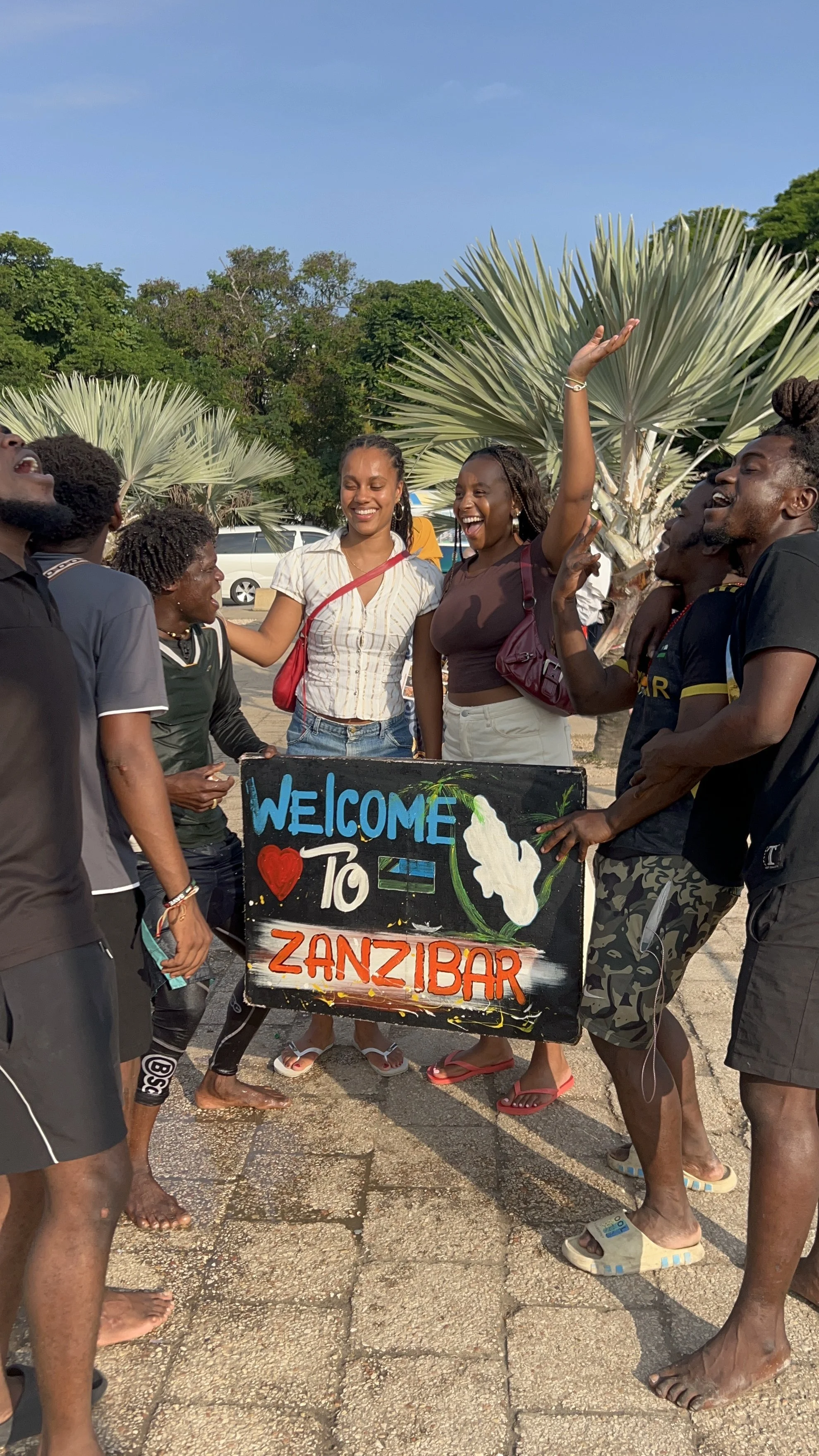 Group of people smiling and standing around a sign that reads 'Welcome to Zanzibar,' outdoors with greenery in the background.