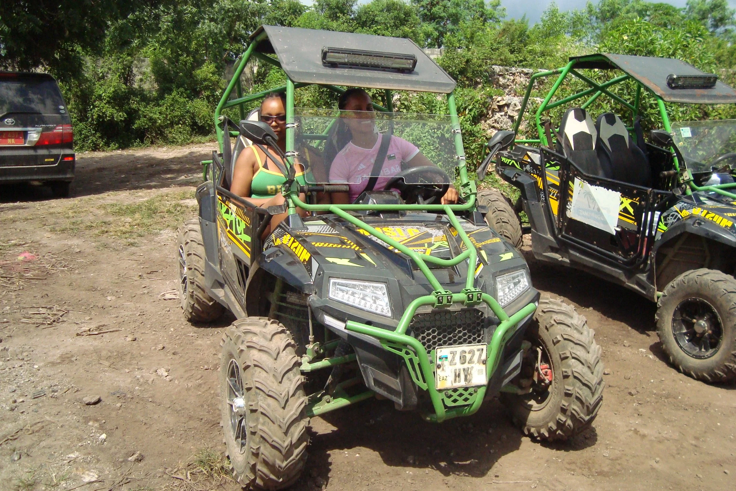 Two women sitting in an off-road utility vehicle, with another similar vehicle parked beside them, in a rural outdoor setting with trees and dirt ground.