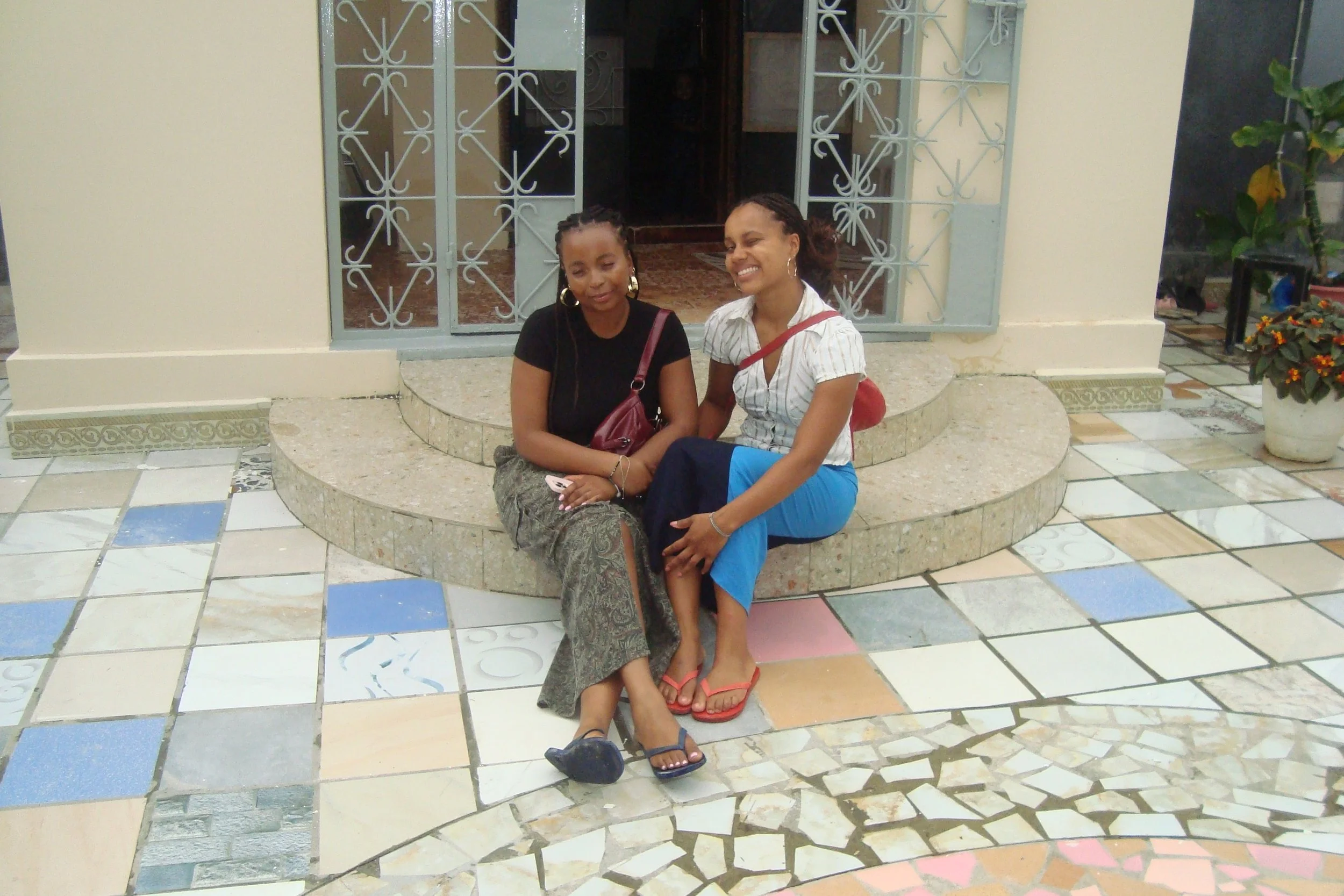 Two women sitting on a stone step outside a building, smiling and posing for the photo. One is wearing a black top and patterned pants, the other is wearing a white striped shirt and blue pants, with both carrying small handbags.