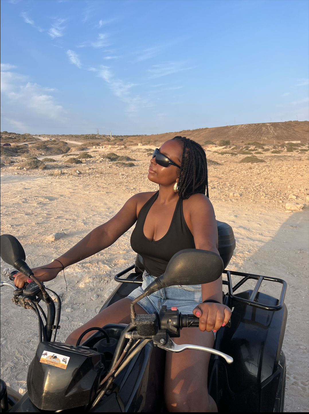 A woman wearing sunglasses, a black tank top, and denim shorts riding a motorcycle in a desert landscape with rocky terrain, sparse bushes, and a clear blue sky.