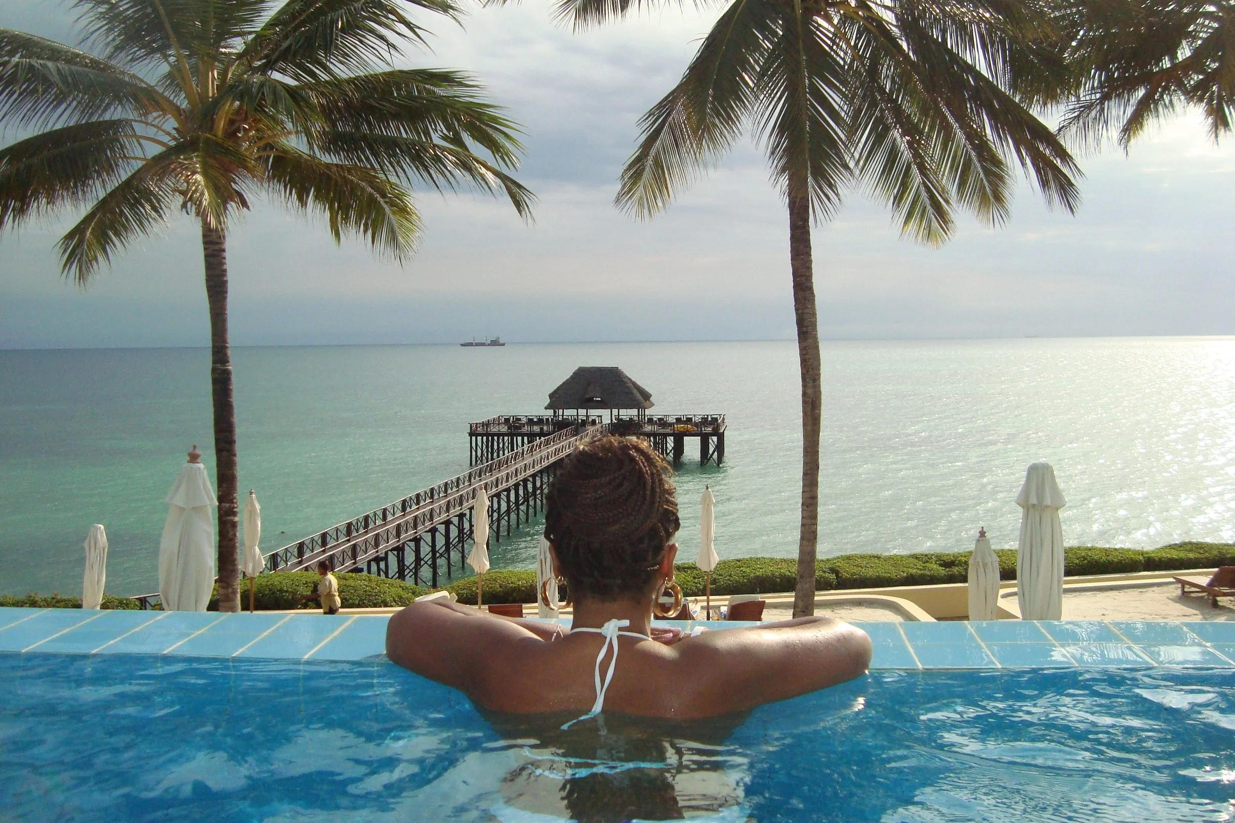 A woman relaxing in a blue pool overlooking the ocean, with palm trees, a pier, and a thatched-roof hut in the background.