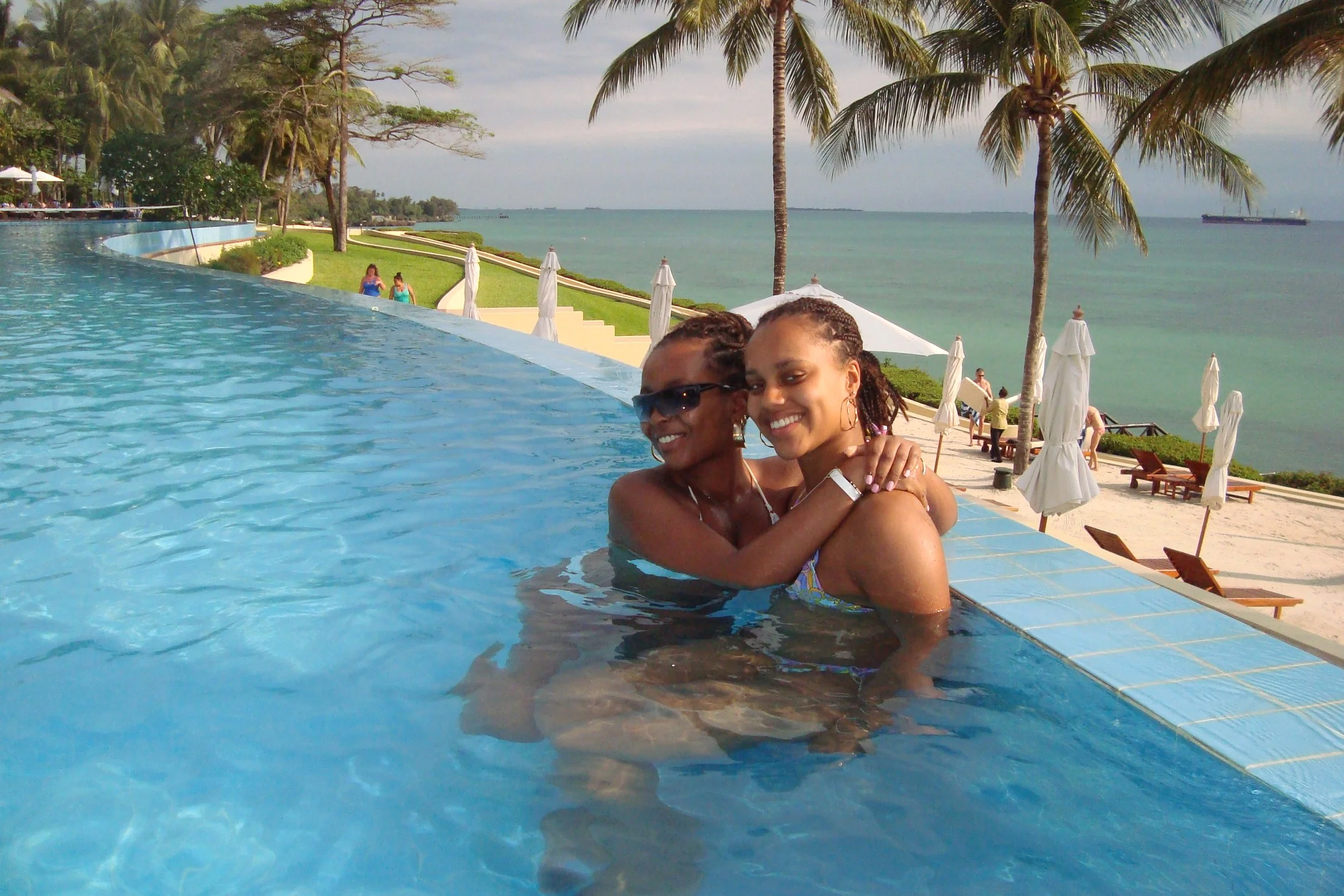 Two women in swimsuits hugging in a swimming pool at a beachside resort with palm trees, lounge chairs, and umbrellas in the background.