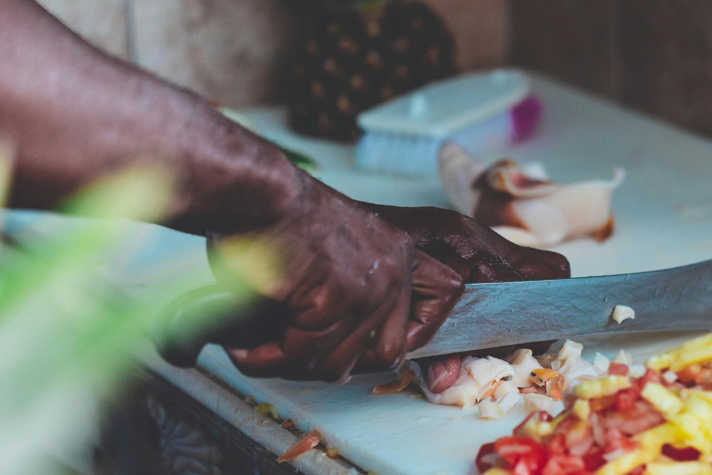 Close-up of a person with dark skin chopping raw chicken on a white cutting board surrounded by chopped vegetables.