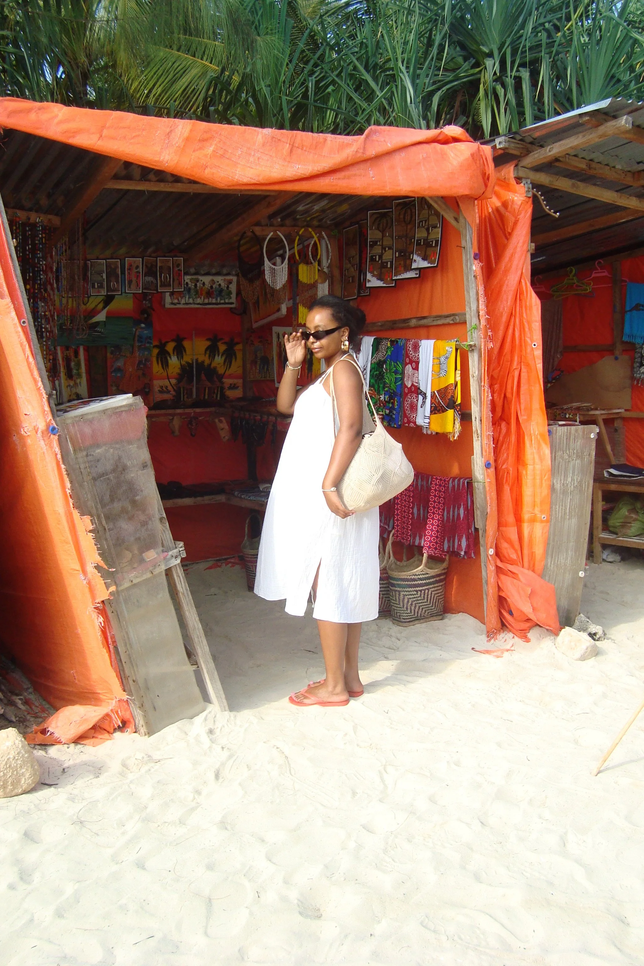 A woman in a white dress and sunglasses standing in front of a beachside vendor stall with colorful textiles and souvenirs, sandy ground, greenery overhead.