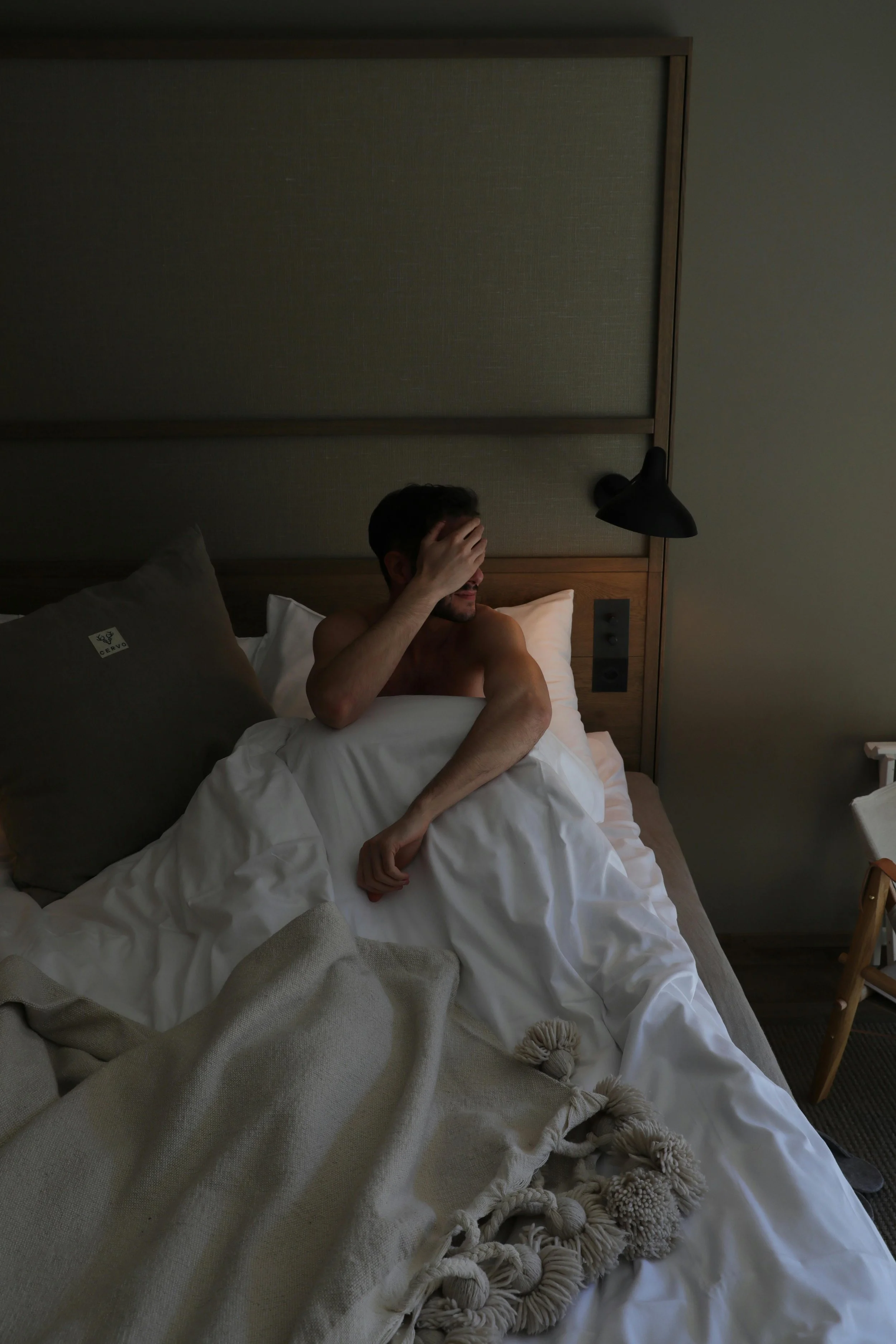 Un homme non habillé, assis dans un lit dans une chambre sombre, se couvrant le visage avec une main.