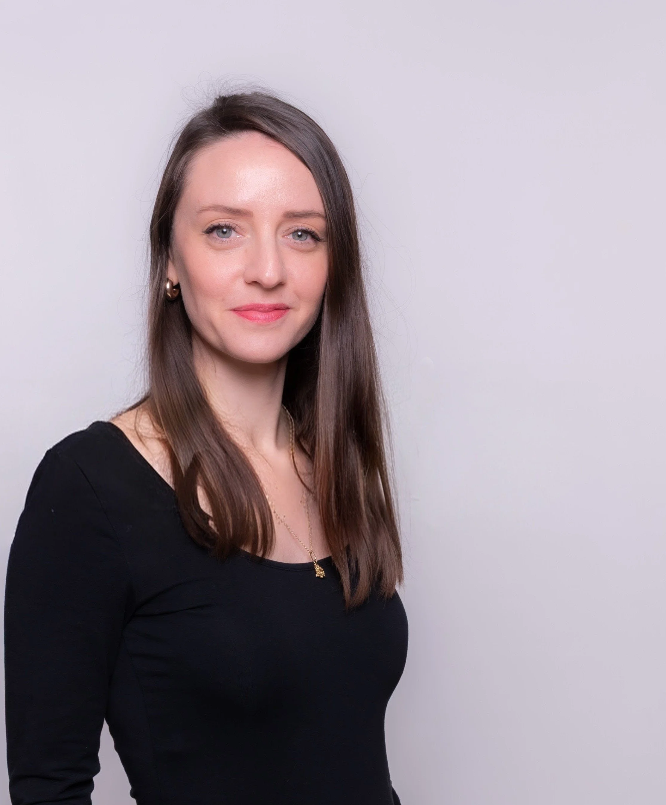 Portrait d'une femme avec des cheveux bruns, portant un col noir, des boucles d'oreilles en or et un collier, souriant légèrement, contre un fond blanc.
