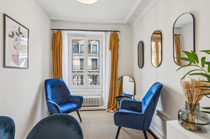 Salon avec deux fauteuils bleus, un miroir, des rideaux jaunes et des murs blancs avec plusieurs miroirs rectangulaires