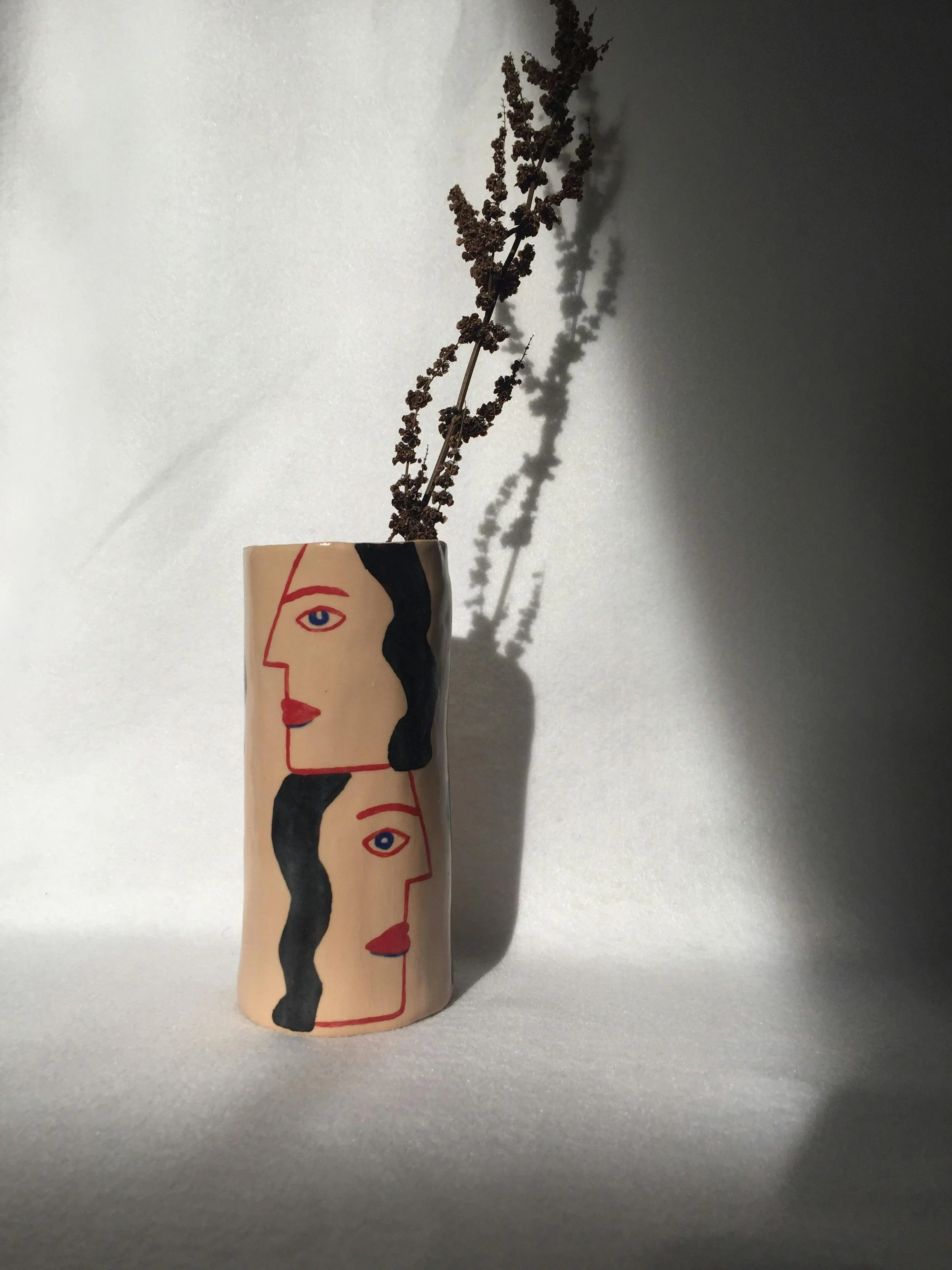 Vase décoré avec un dessin stylisé de visages féminins, contenant une branche de lavande sèche, sur fond neutre avec ombre portée.