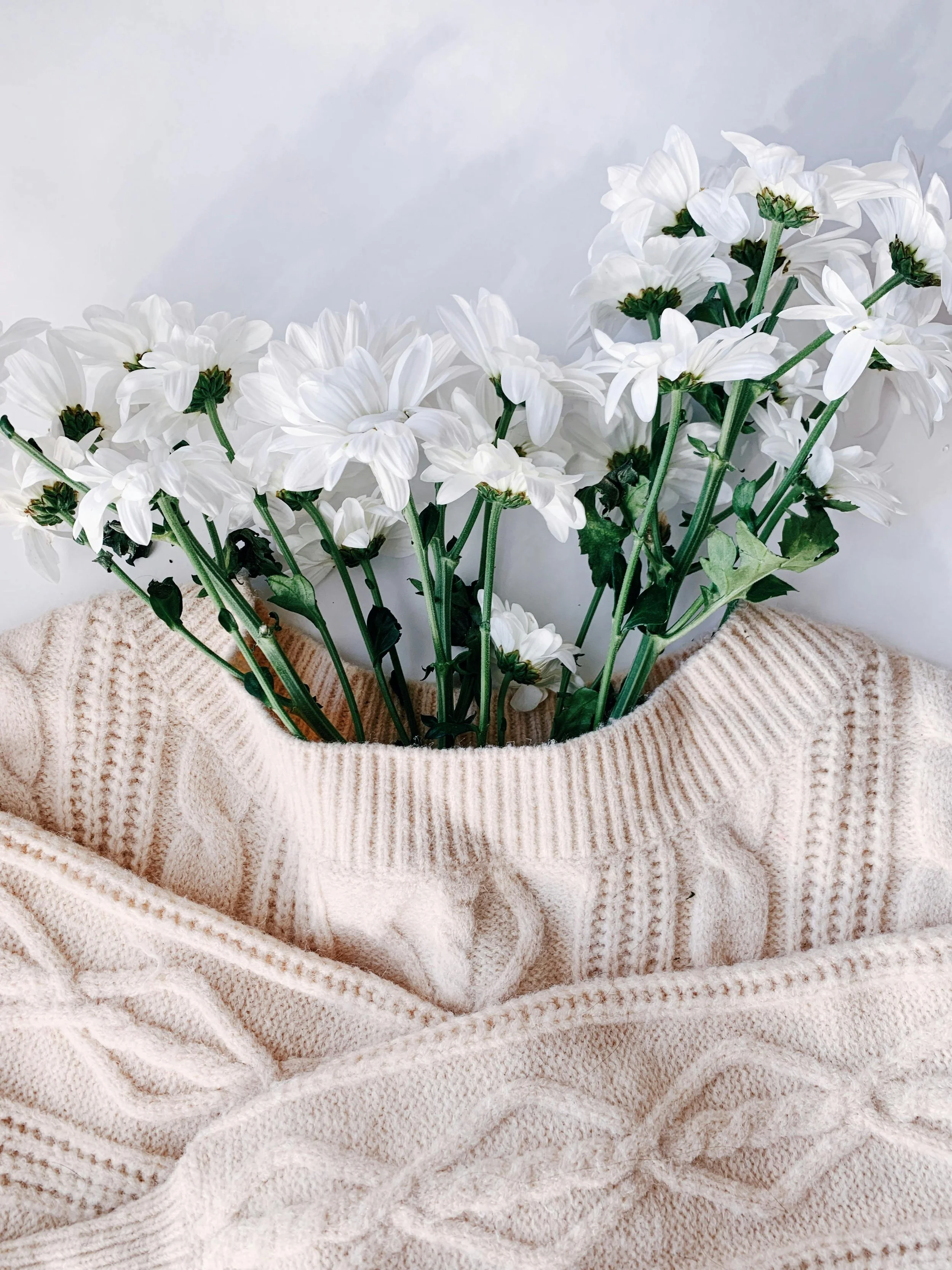 Fleurs blanches dans un vase posé sur un pull en laine beige.