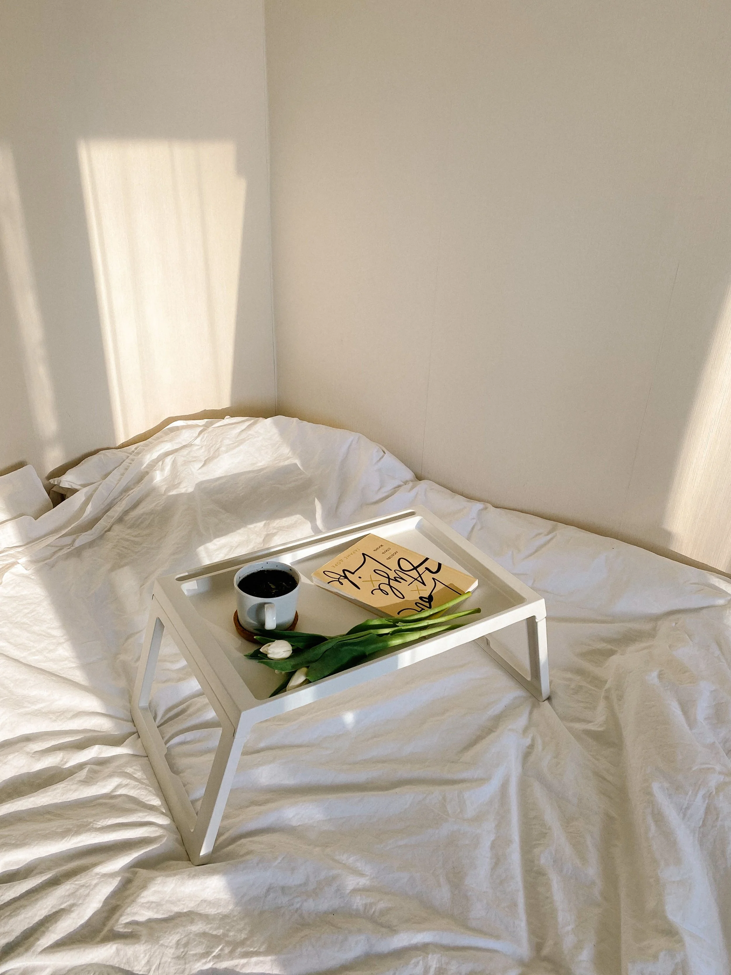 Un plateau blanc avec une tasse de café noir, un livre intitulé 'Love, Shine & Grow', et un bouquet de tulipes blanches sur un lit aux draps blancs dans une chambre ensoleillée.