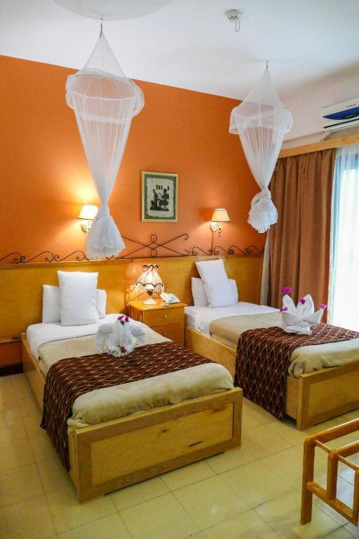 A hotel room with two twin beds, each decorated with folded towels and flowers. There are hanging mosquito nets above the beds, a nightstand with a lamp between the beds, and wall-mounted lamps above them. The room has a framed picture on an orange accent wall, and beige curtains covering a window or sliding door with sunlight coming through.