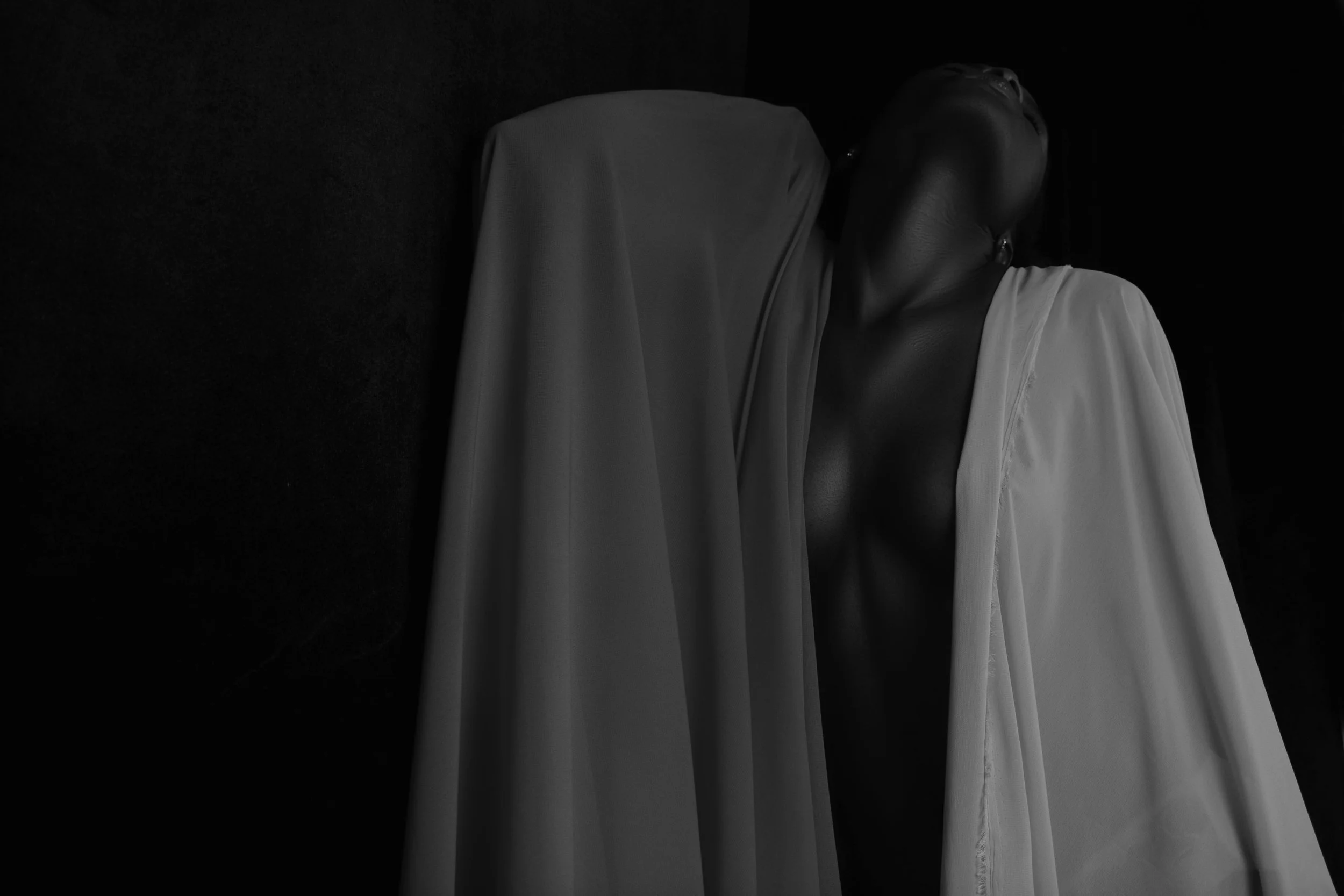 A woman leaning against a wall with her eyes closed, wearing a white dress and a necklace, with her head and shoulders visible in a dark room.