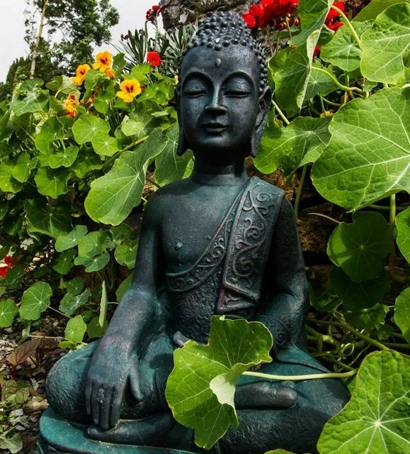 A Buddha statue sitting in a garden surrounded by green ivy and colorful flowers.