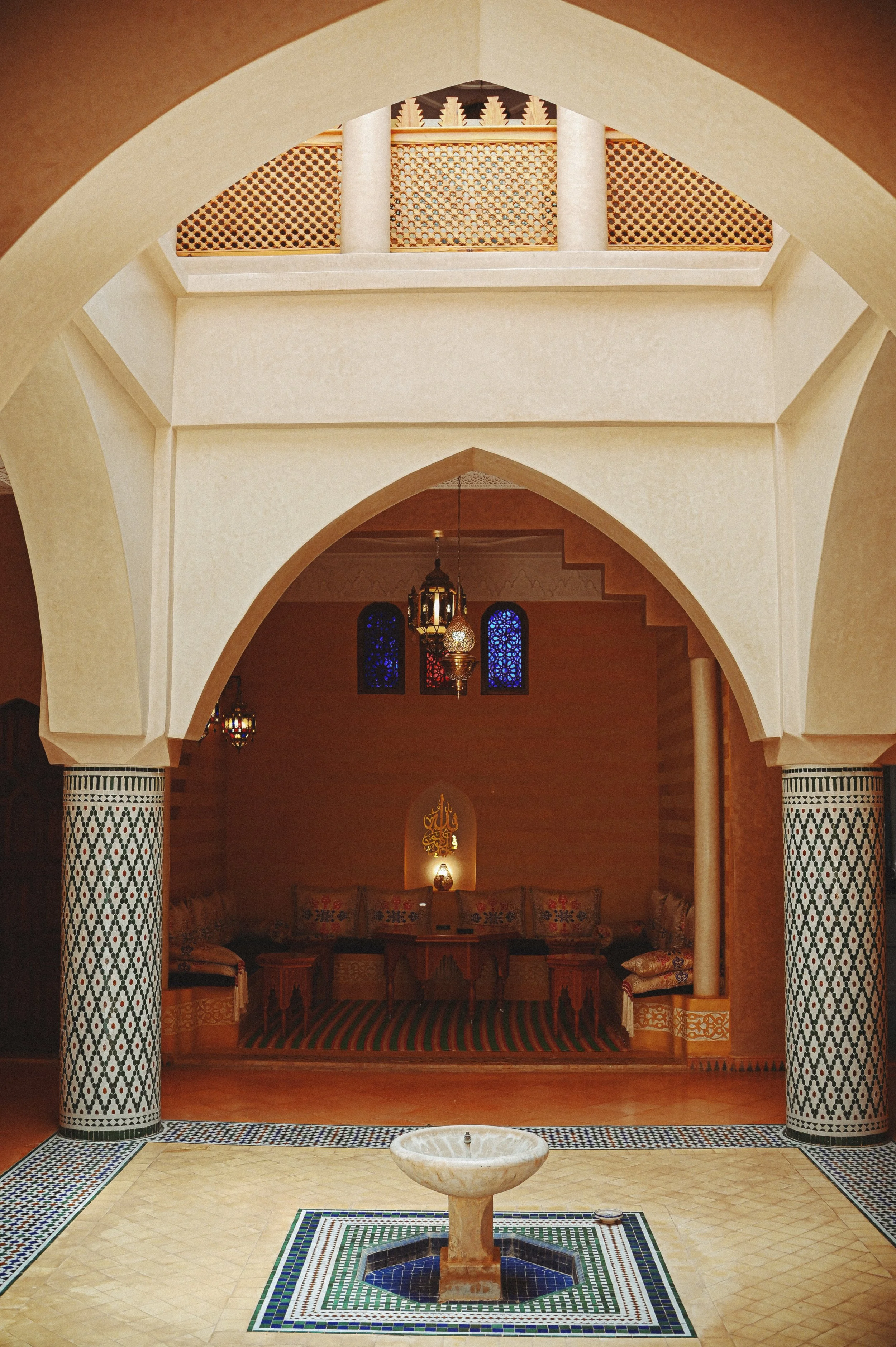 Interior of a mosque with arches, columns with intricate tile designs, hanging lamps, a fountain in the center, and a prayer area with cushions and a statue or symbol in the background.