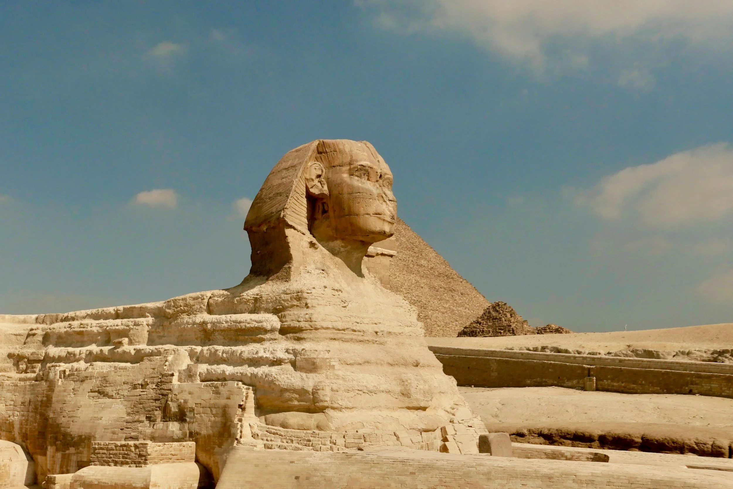 Ancient Egyptian Sphinx statue with a pyramid in the background, under a blue sky with scattered clouds.