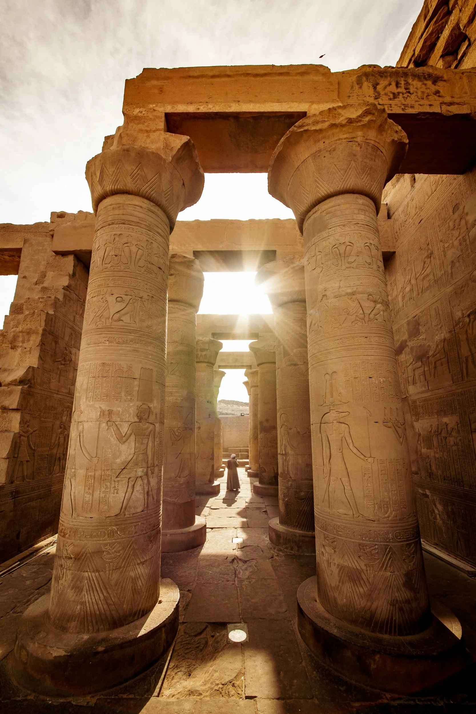 Ancient Egyptian temple interior with large stone columns decorated with hieroglyphics and carvings, sunlight streaming through openings at the top, and a person walking in the background.