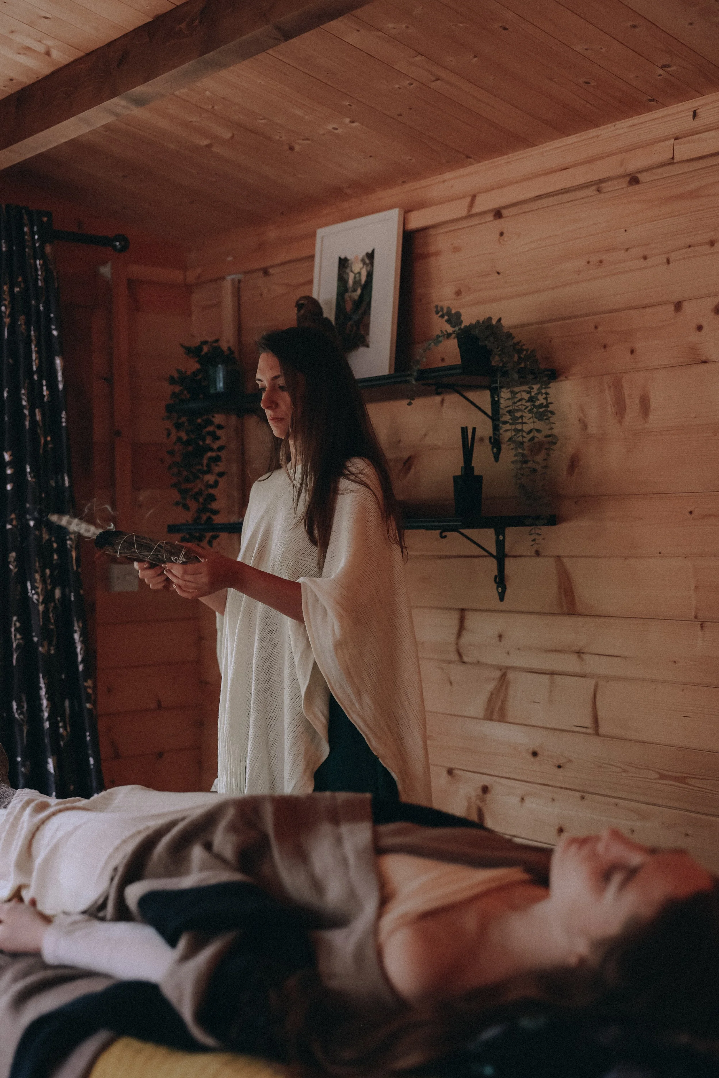 A woman with long dark hair wearing a white top is standing indoors near a wooden wall, looking at her phone. The room has wooden paneling, a framed picture, and plants on shelves. In the foreground, a dog with black, white, and brown fur is lying on a bed or couch.