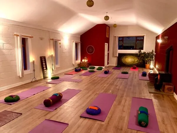A yoga studio with purple mats and bolsters arranged on a wooden floor, illuminated by warm wall lights, with windows and potted plants in the background.