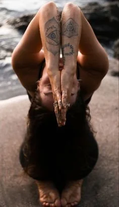 Person with tattoos on arms and fingers performing a yoga pose on a rocky surface near water, with hair hanging downward.