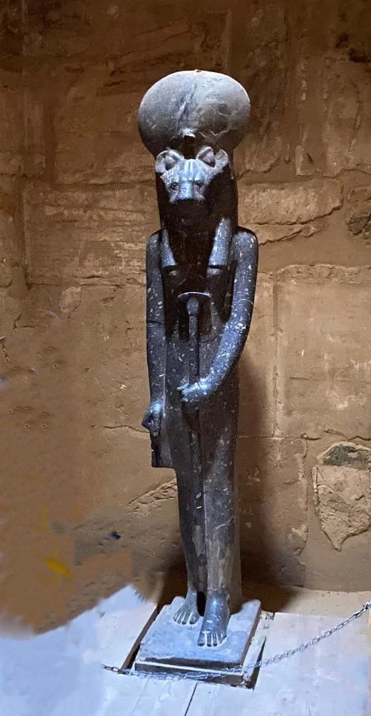 A dark-colored statue of a person with an oversized skull, standing on a small platform against a stone wall, with arms bent and hands holding a rod or staff.
