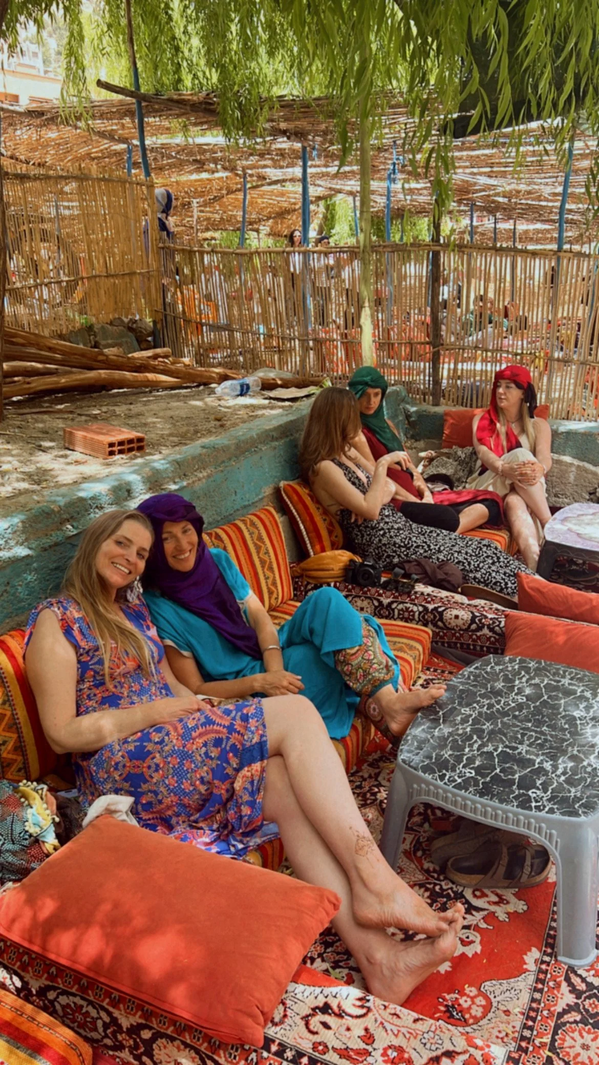 Group of women sitting on colorful cushions and chairs, engaged in conversation, in an outdoor setting with bamboo fencing and a shaded area overhead.