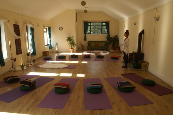 A yoga studio with purple mats and folded blankets arranged in a circle, a woman standing by a television, and a window with green curtains letting in natural light.