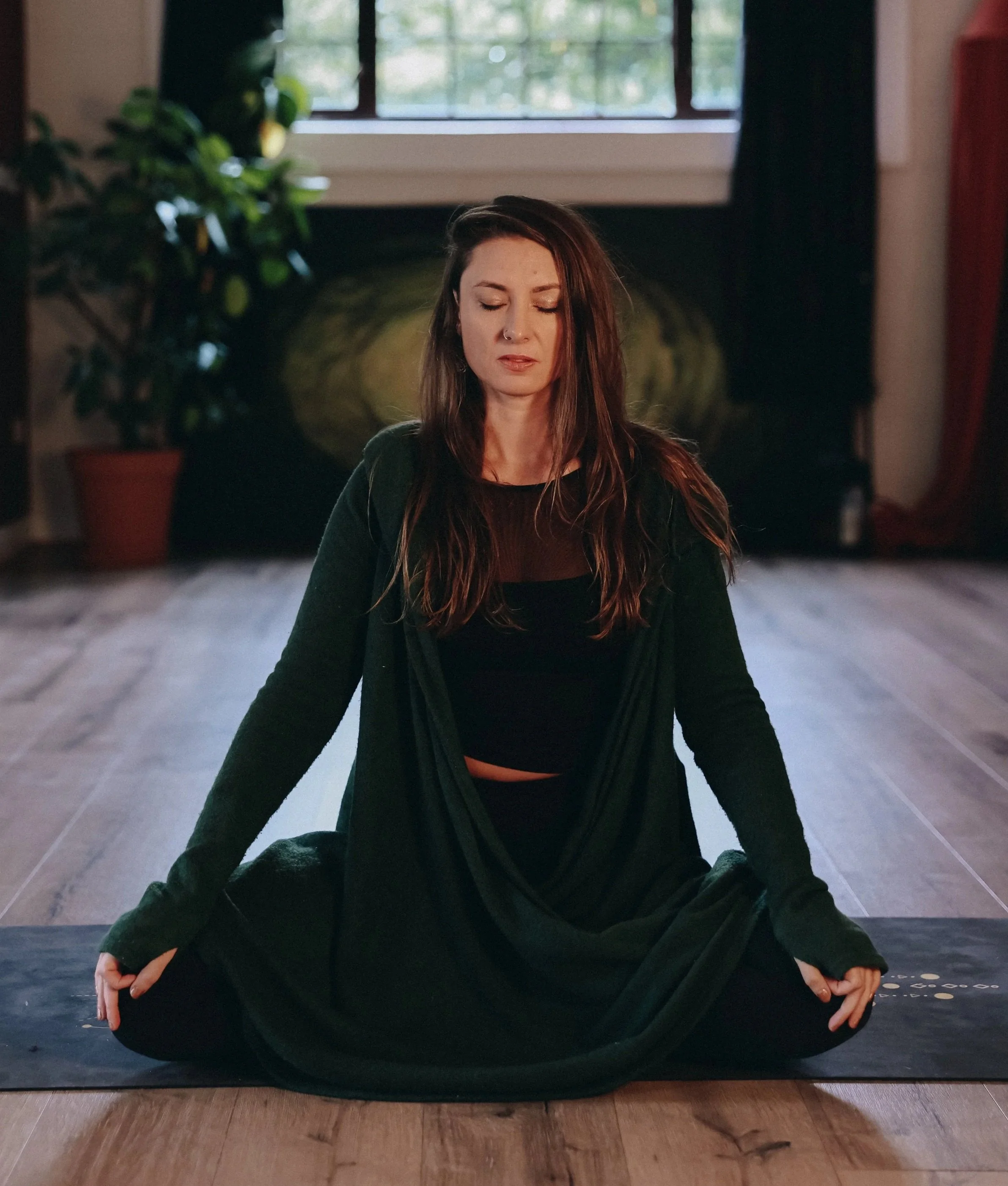 A woman practicing yoga in a seated cross-legged position on a yoga mat in a room with a window and potted plant.