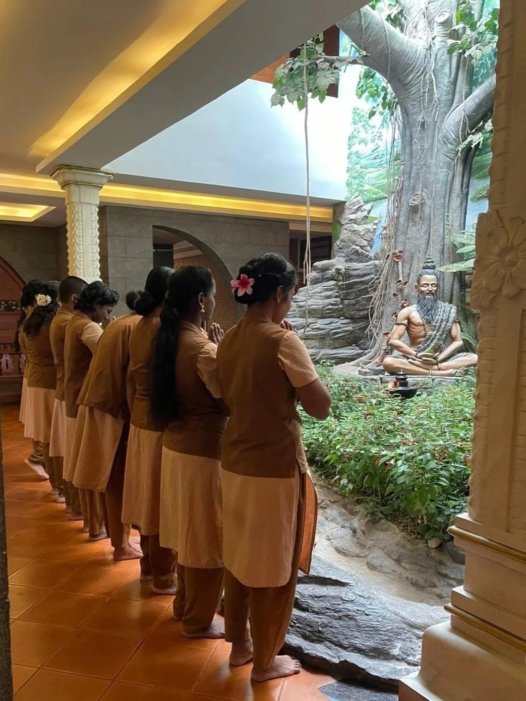 A group of women dressed in traditional brown clothes with flowers in their hair are praying indoors in front of a garden scene with a large statue of a meditative sage next to a tall tree with hanging roots.