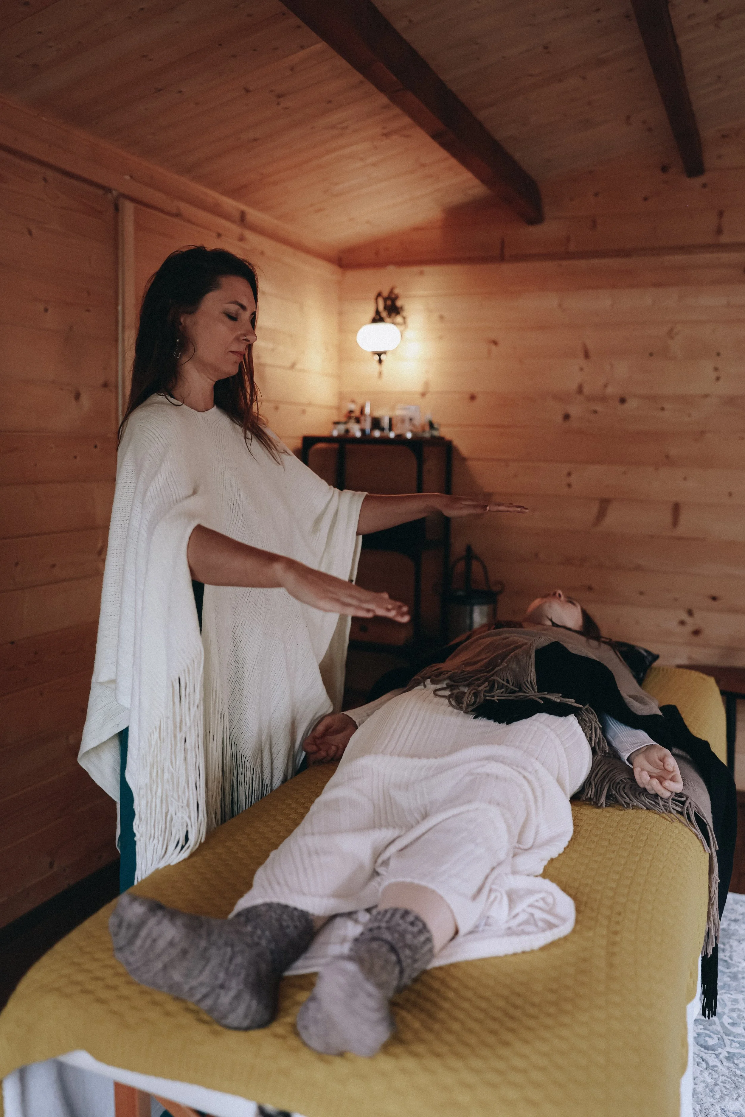 A woman with long dark hair making a gesture over a person lying on a massage table in a wooden room.