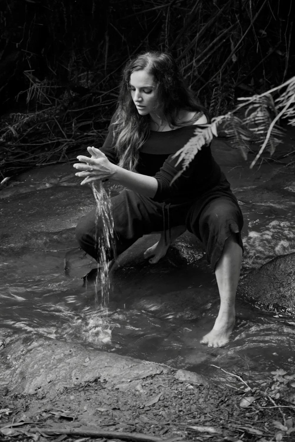 A woman in a long-sleeve dress is sitting on a rock in a creek, with her feet and hands in the water. She is surrounded by dense foliage and appears to be splashing or playing with the water.