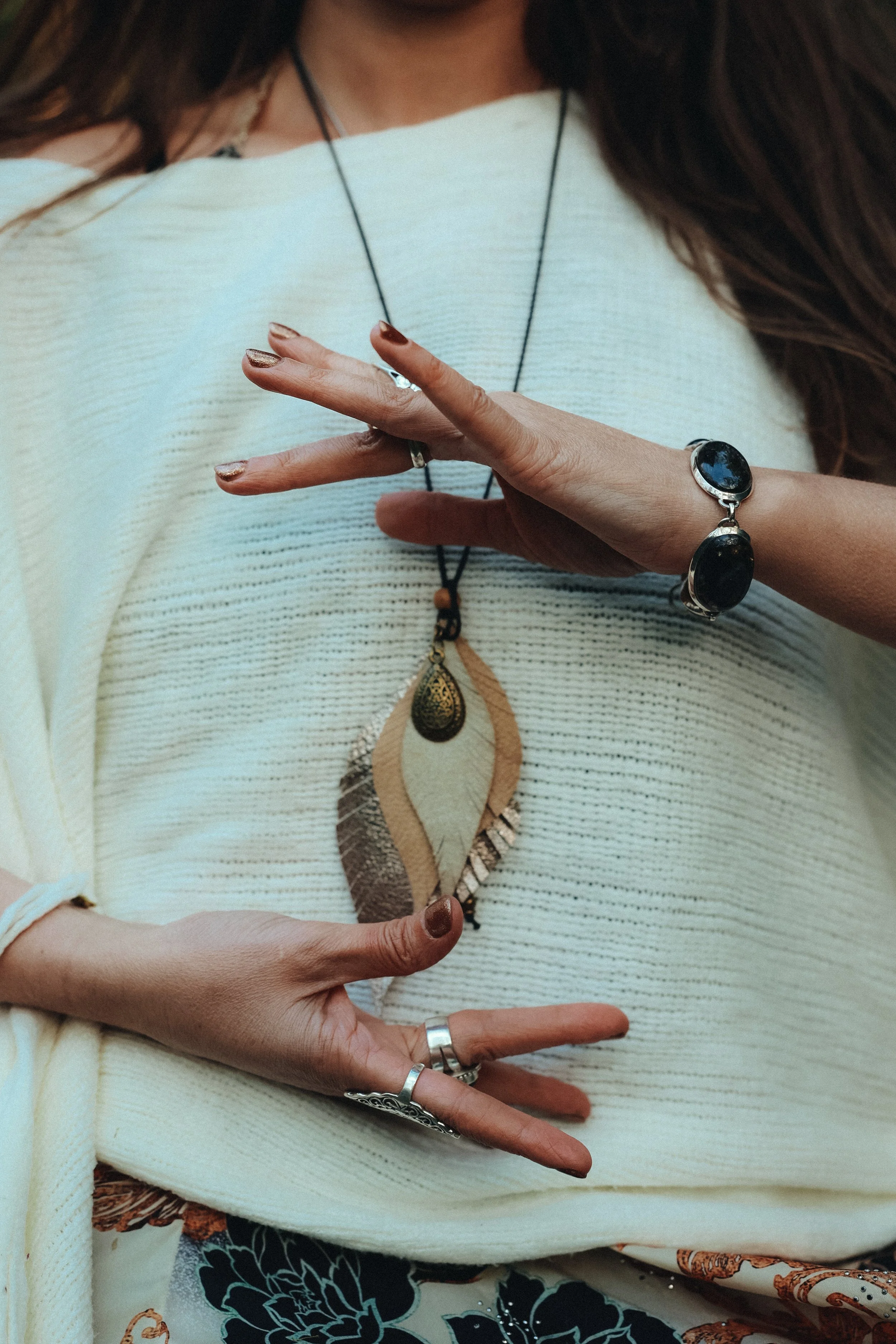 Close-up of a woman's hands and upper torso, adorned with jewelry including rings, bracelets, and necklaces, while wearing a light-colored sweater.