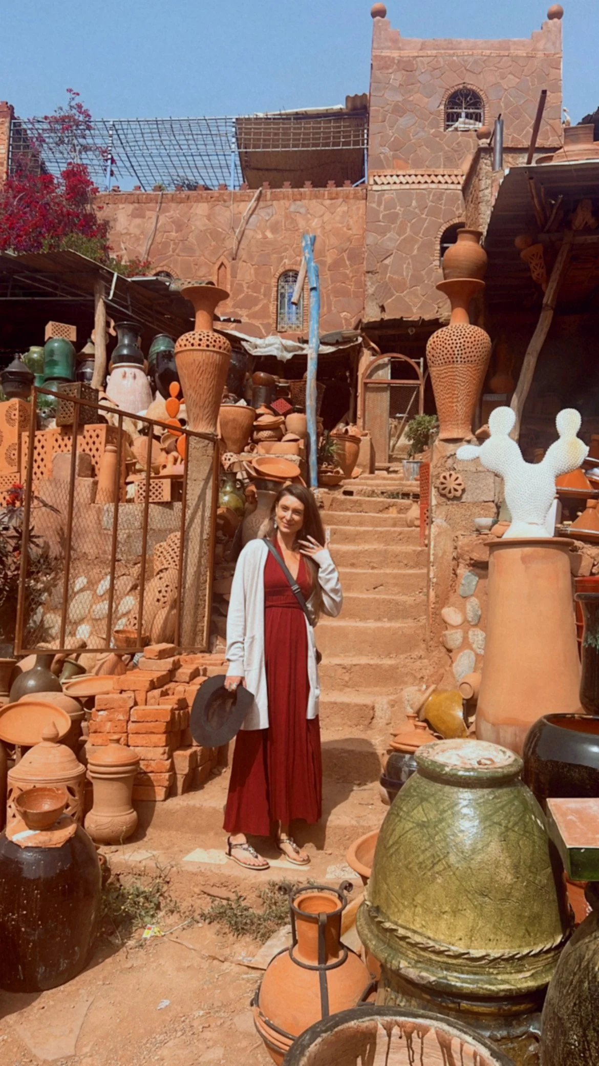 A woman standing in front of a rustic outdoor pottery shop with stairs, terracotta pots, and a stone building in the background.