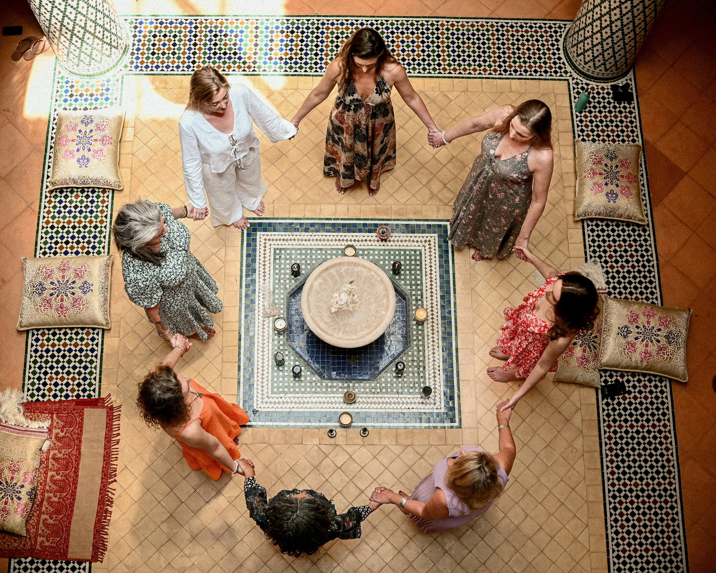 Six women and two men standing in a circle holding hands in a room with Moroccan-style tile flooring and walls, pillows on the ground, some candles on a table, and shoes and water bottles nearby.