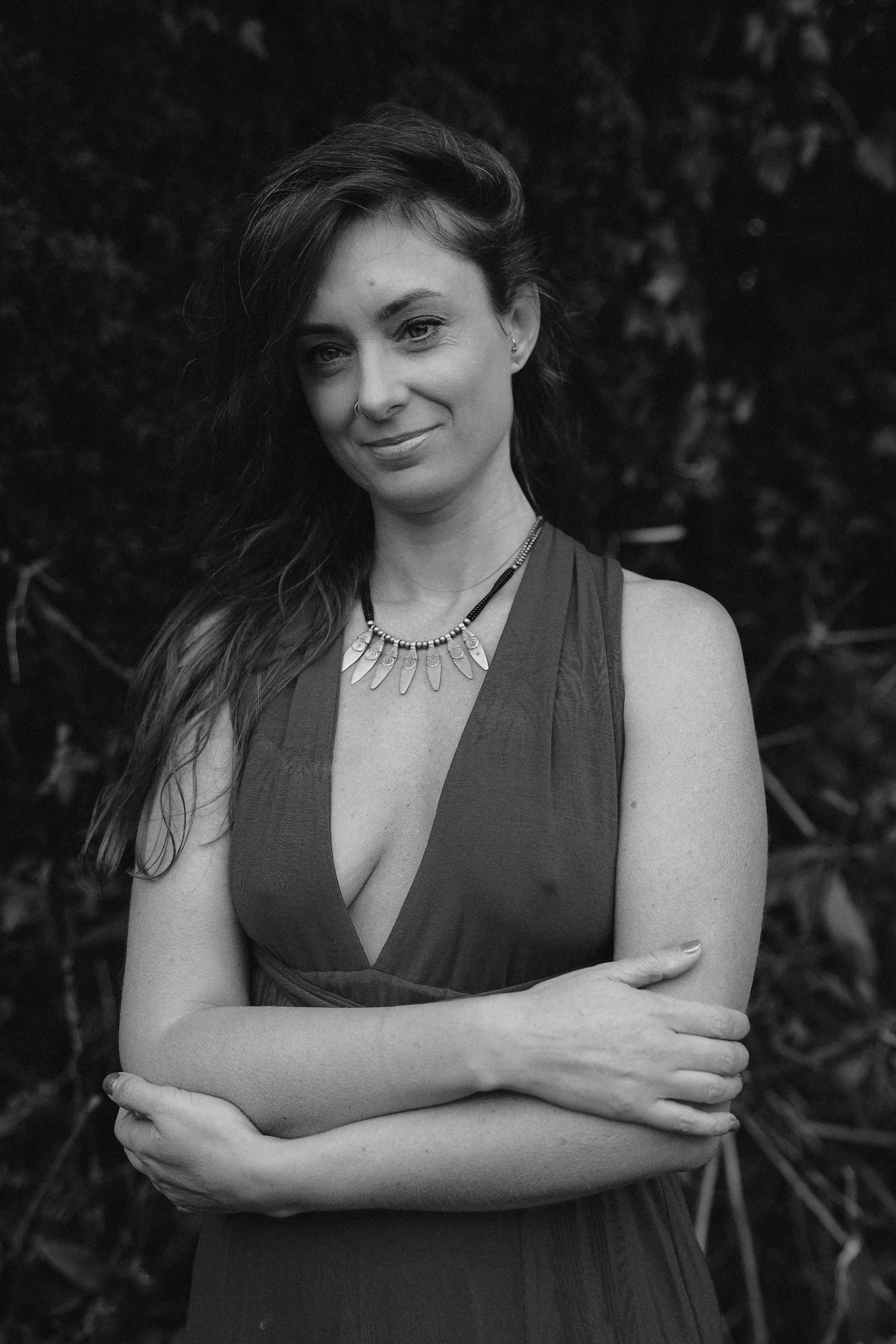 Black and white portrait of a woman with long hair, wearing a sleeveless dress and a necklace, standing outdoors against a blurred background of foliage.