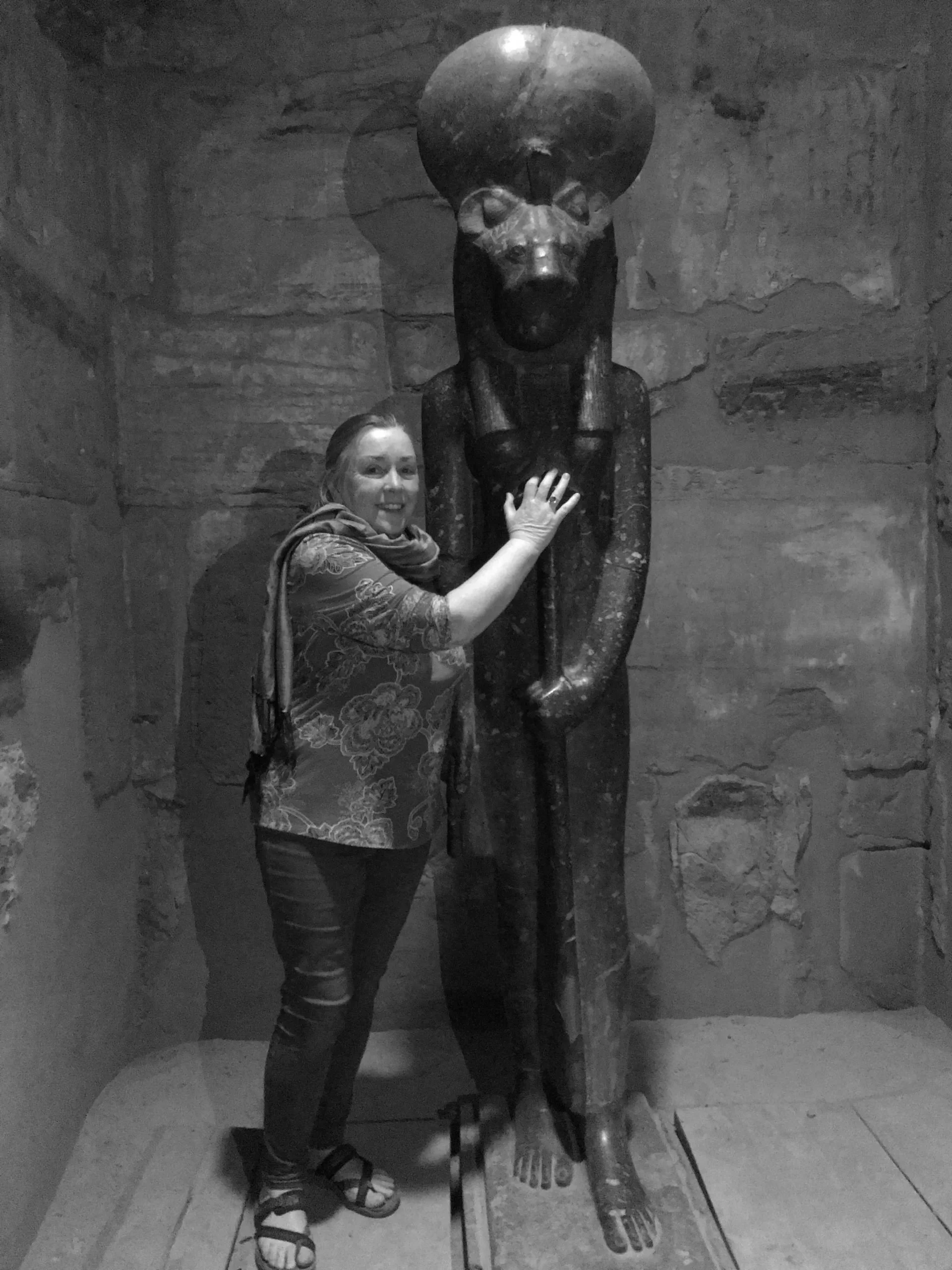 A woman in casual clothing and sandals standing next to a large ancient Egyptian statue of a seated figure with a human body and a lion head, holding a staff, inside a stone-walled room.
