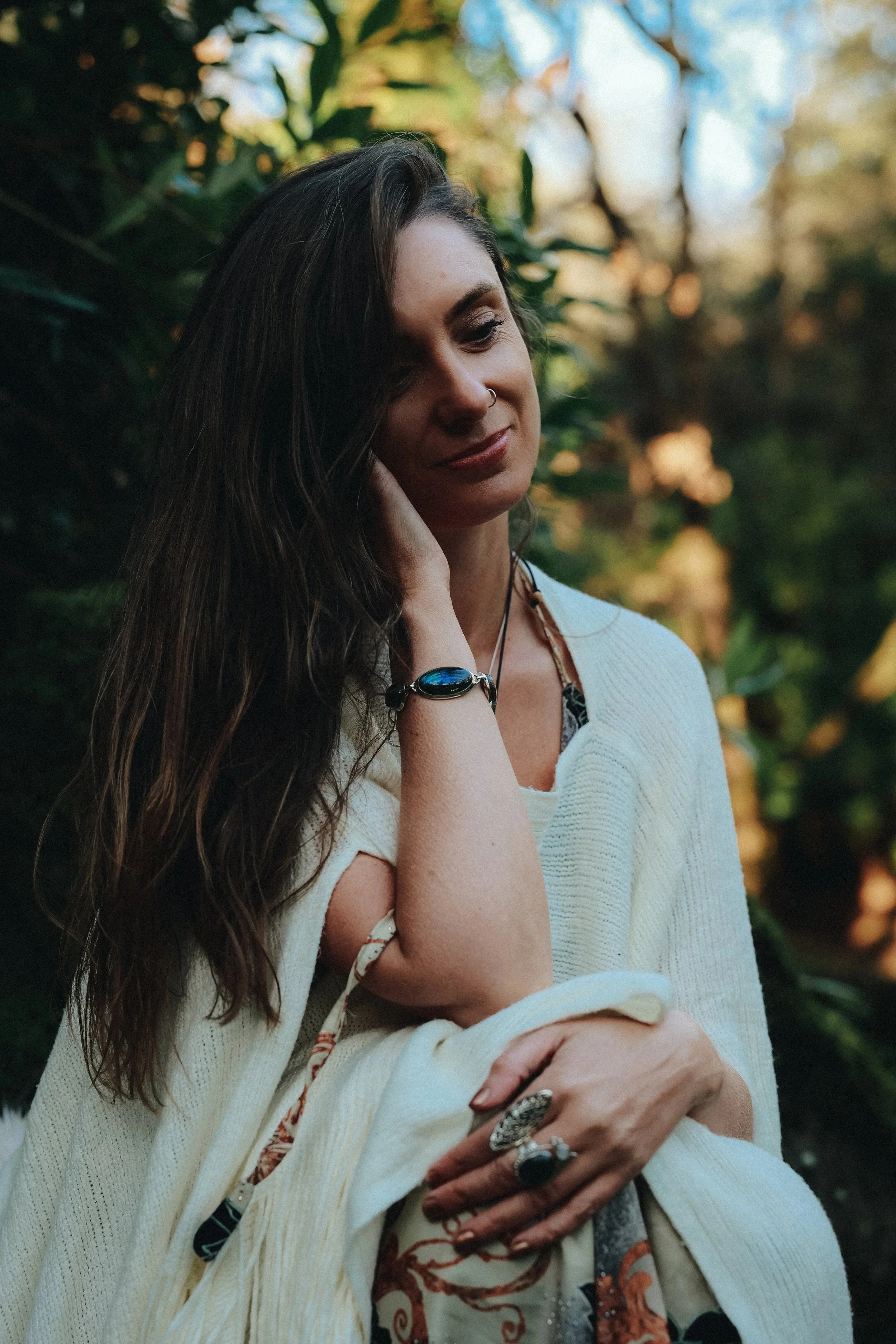 A woman with long dark hair and a nose ring is smiling with her eyes closed. She is wearing a cream-colored cardigan, a ring with a large dark stone, and a dark bracelet. She is touching her face with her left hand and holding her right arm. The background features outdoor foliage with blurred green leaves and sunlight.