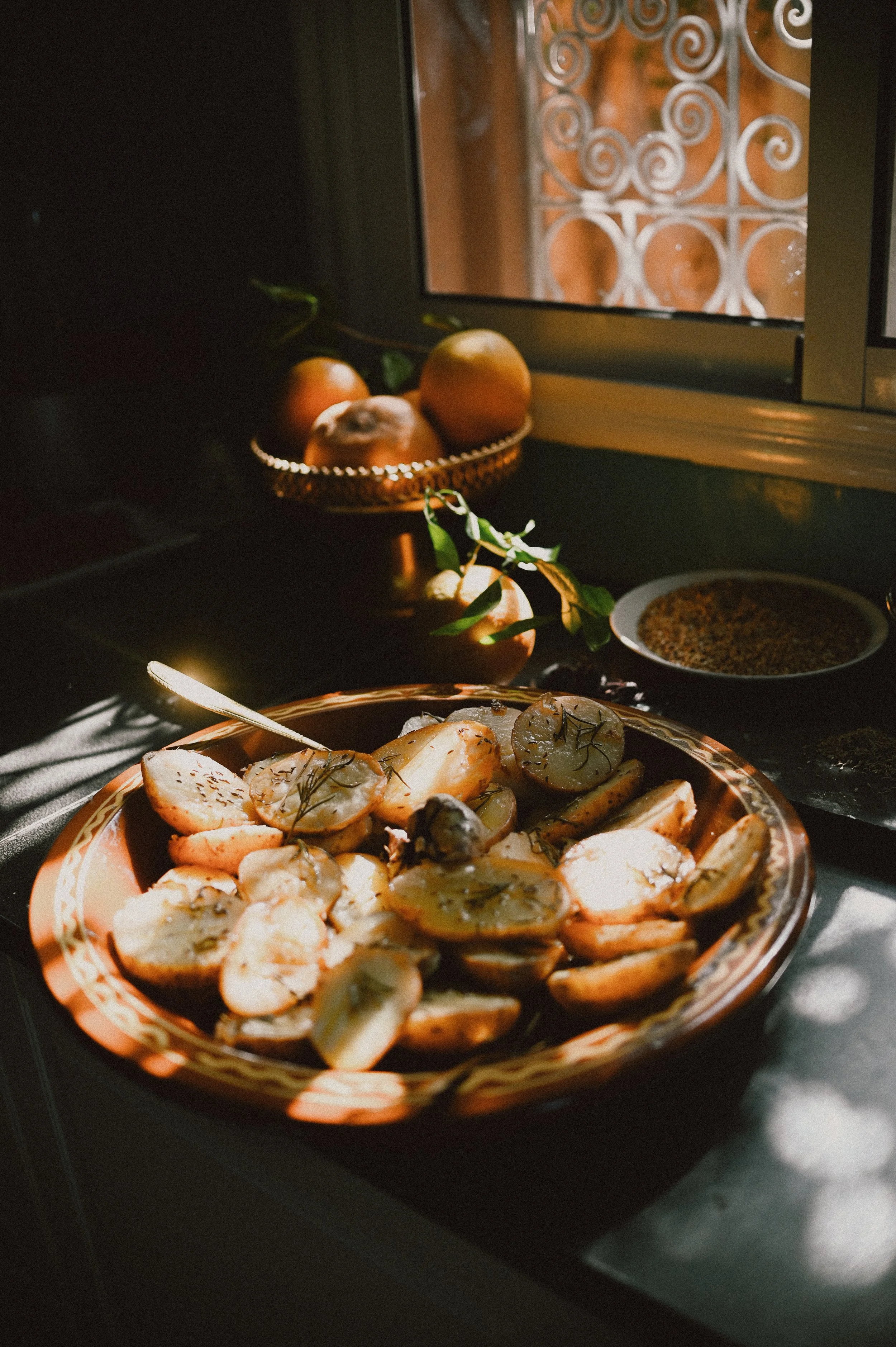 A plate of sliced potatoes garnished with herbs on a table near a window, with a bowl of seasoning and a basket of onions in the background.