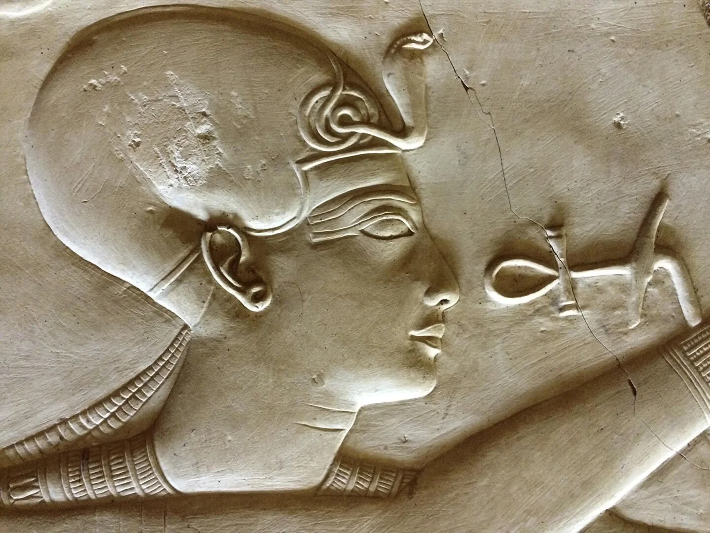Carved stone relief of an Egyptian woman in profile with a headdress and traditional jewelry, facing and touching a small figure of a man.