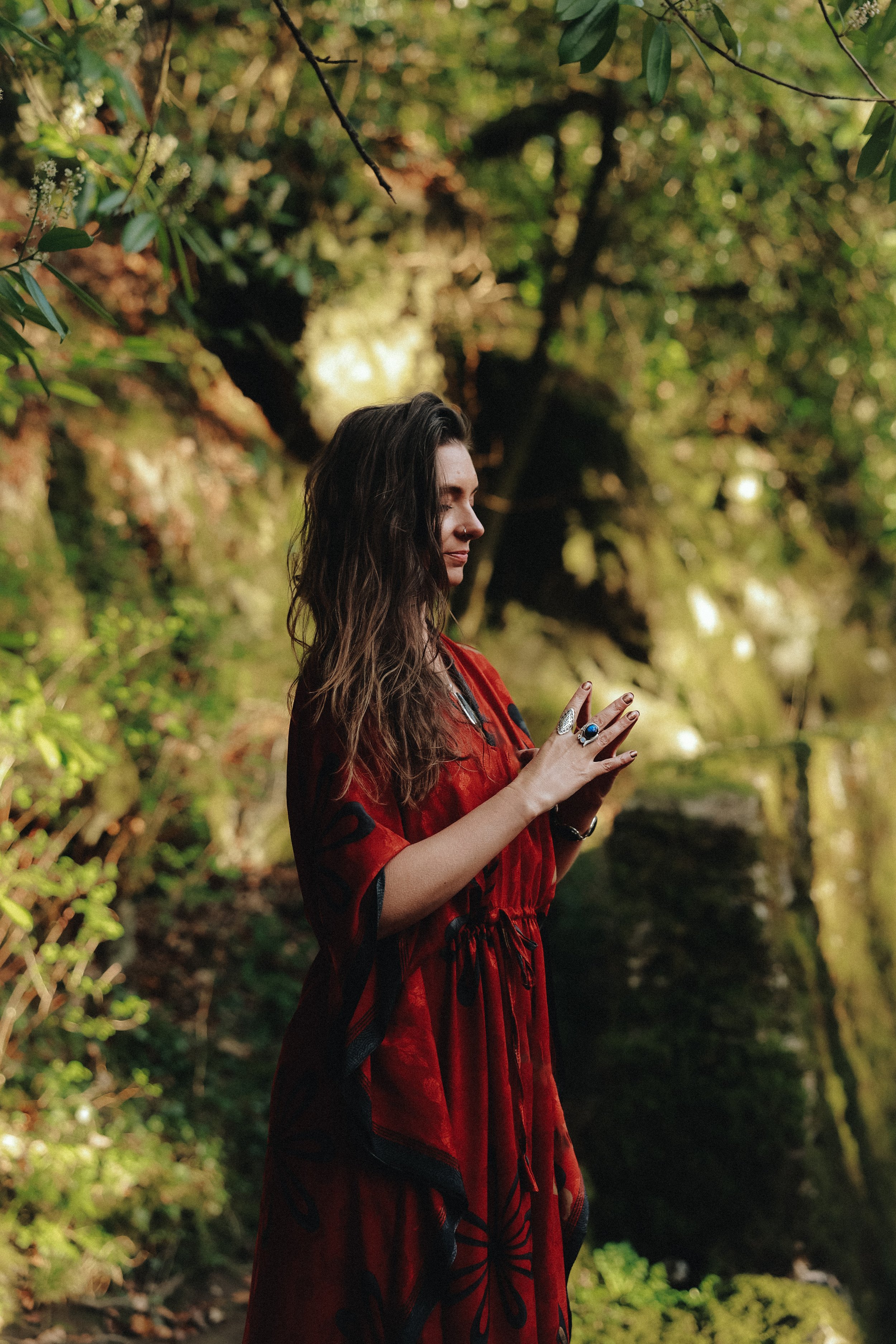 A young woman with long dark hair and tattoos on her hands, wearing a red dress with black patterns, standing outdoors in a green forest, with her eyes closed and hands pressed together in a meditative pose.