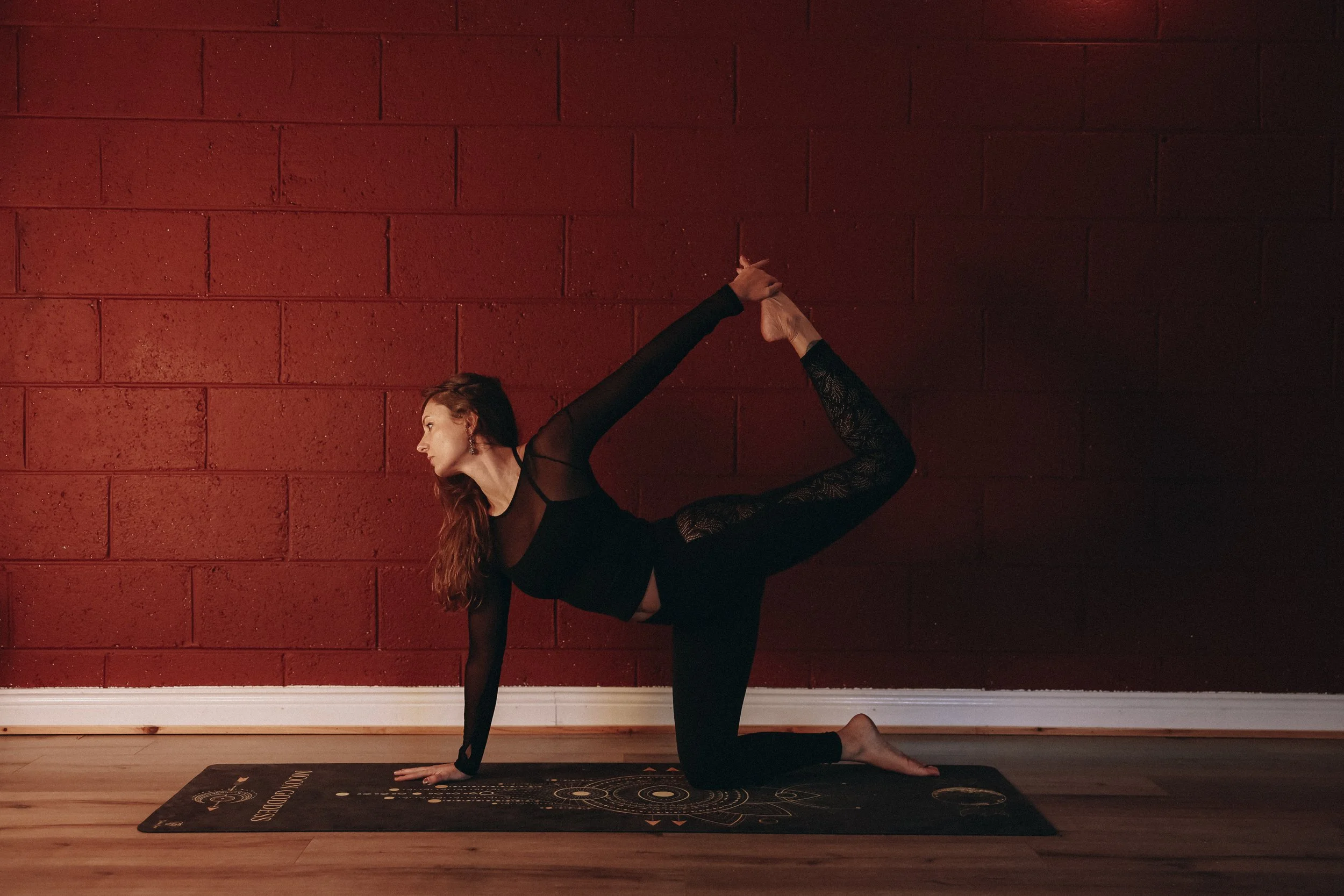 A woman with long red hair practicing yoga indoors against a red brick wall, performing a backbend with one leg extended upward and reaching back to hold her foot, on a black yoga mat.