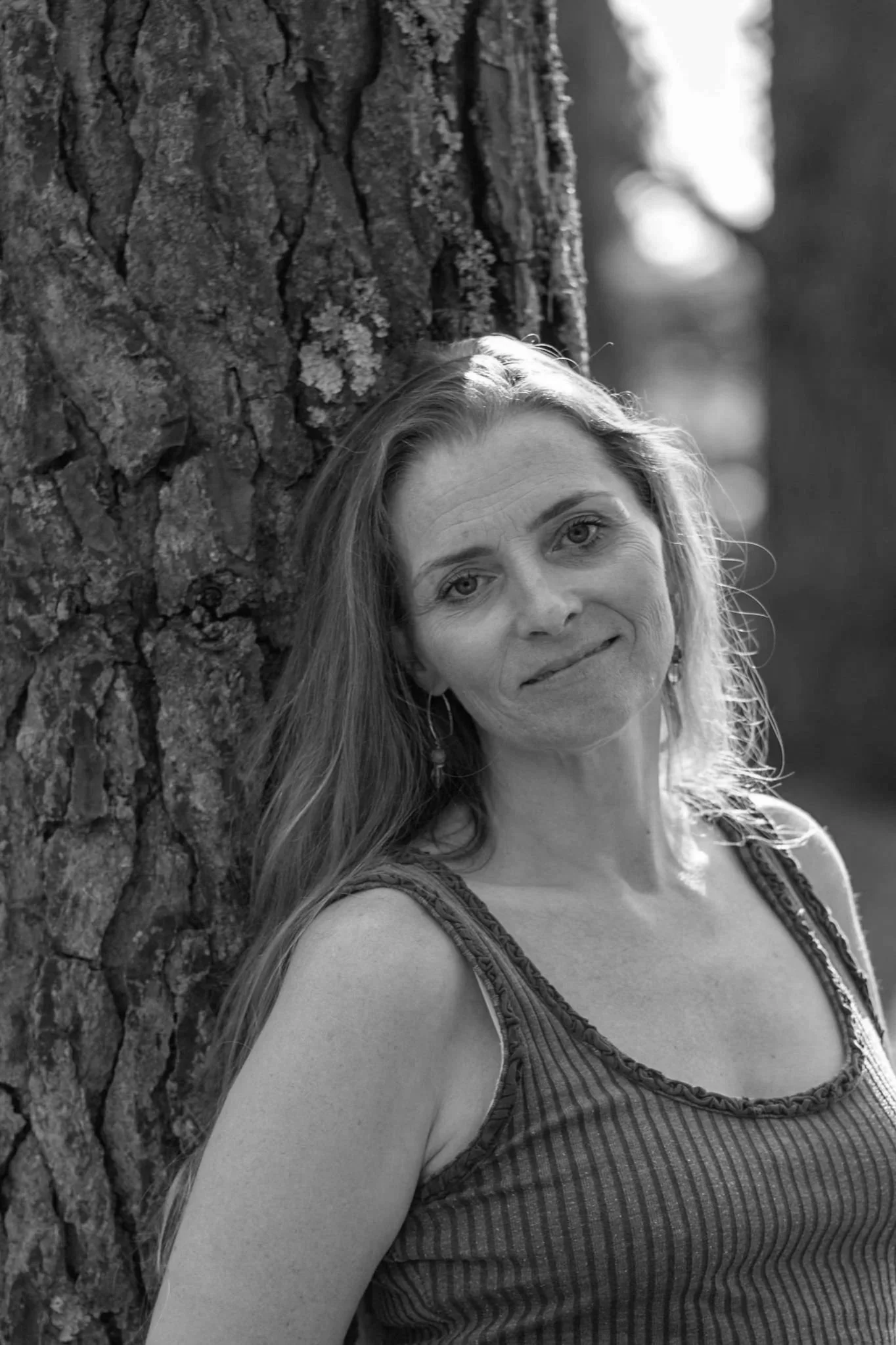 A woman with long hair leaning against a tree trunk in a forest, wearing a striped sleeveless top, smiling slightly, in black and white.